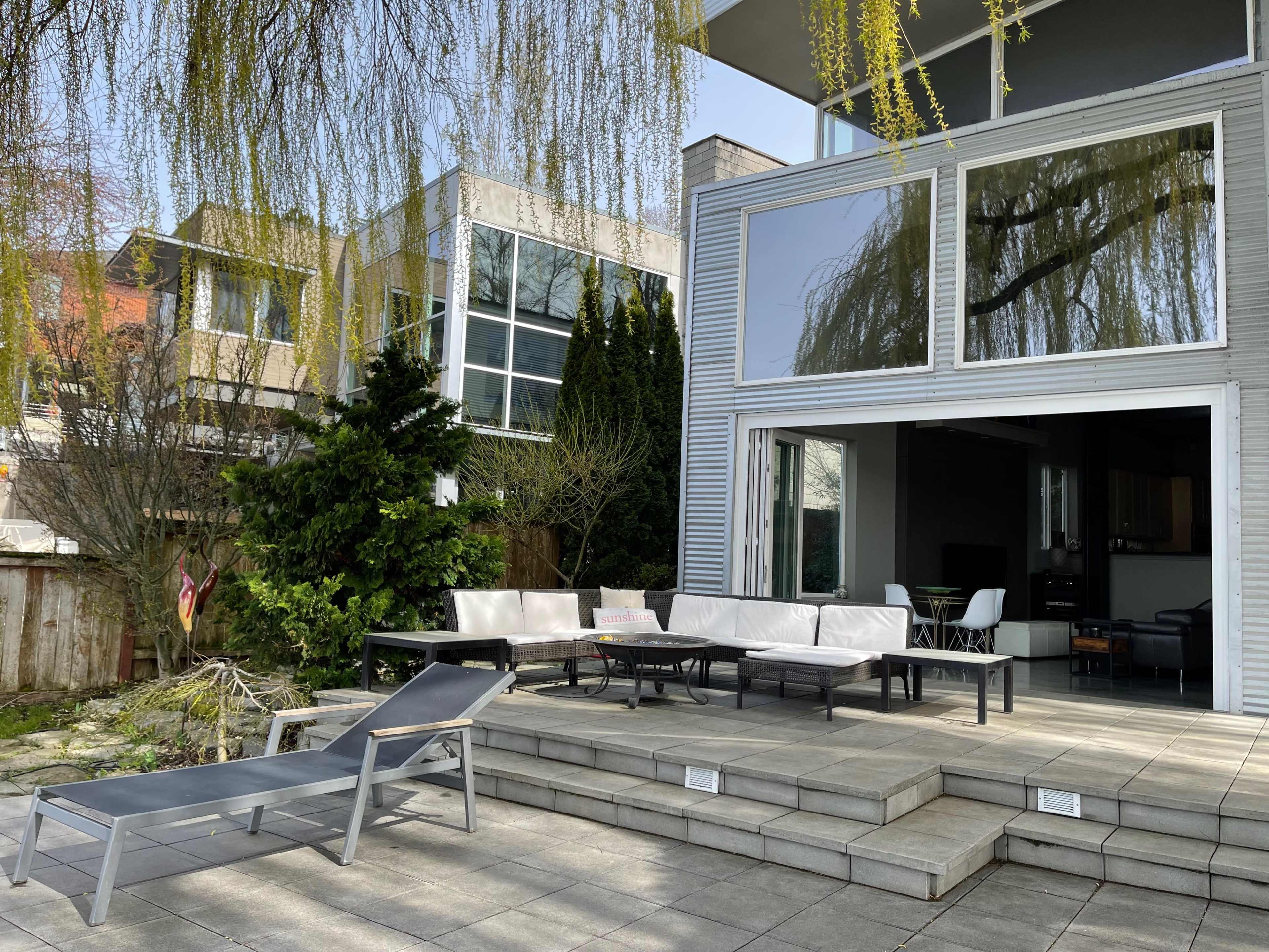 A modern outdoor patio with a seating area, a chaise lounge, and a backdrop of contemporary buildings and trees.