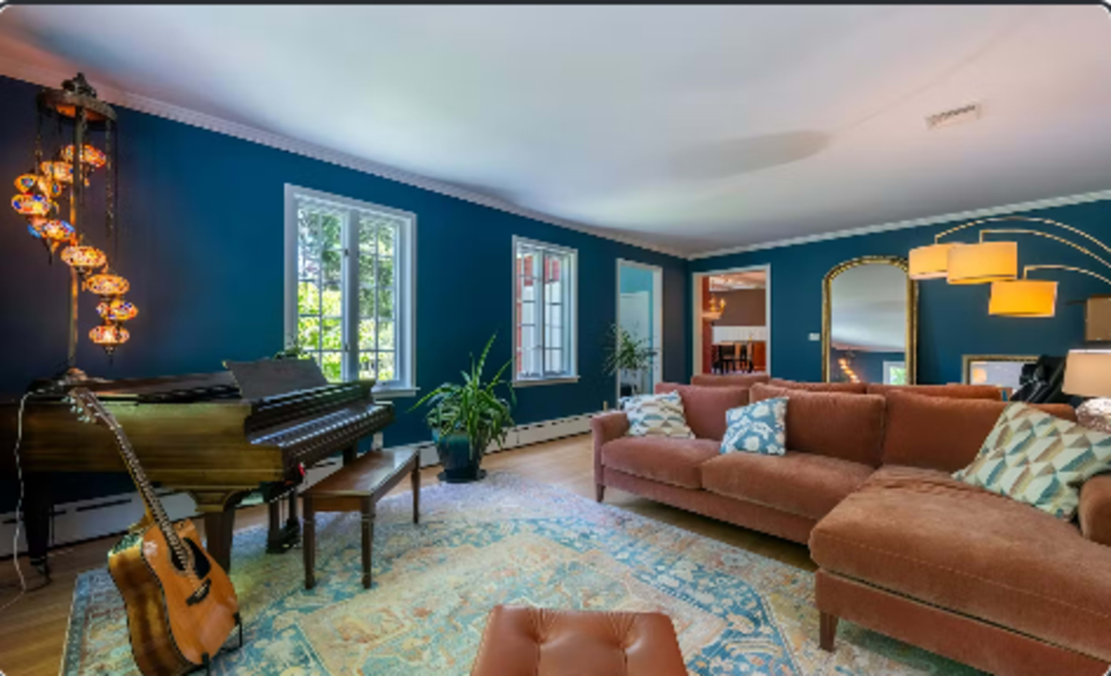 The image shows a living room featuring a brown sofa, a piano, several windows allowing natural light, and decorative plants.
