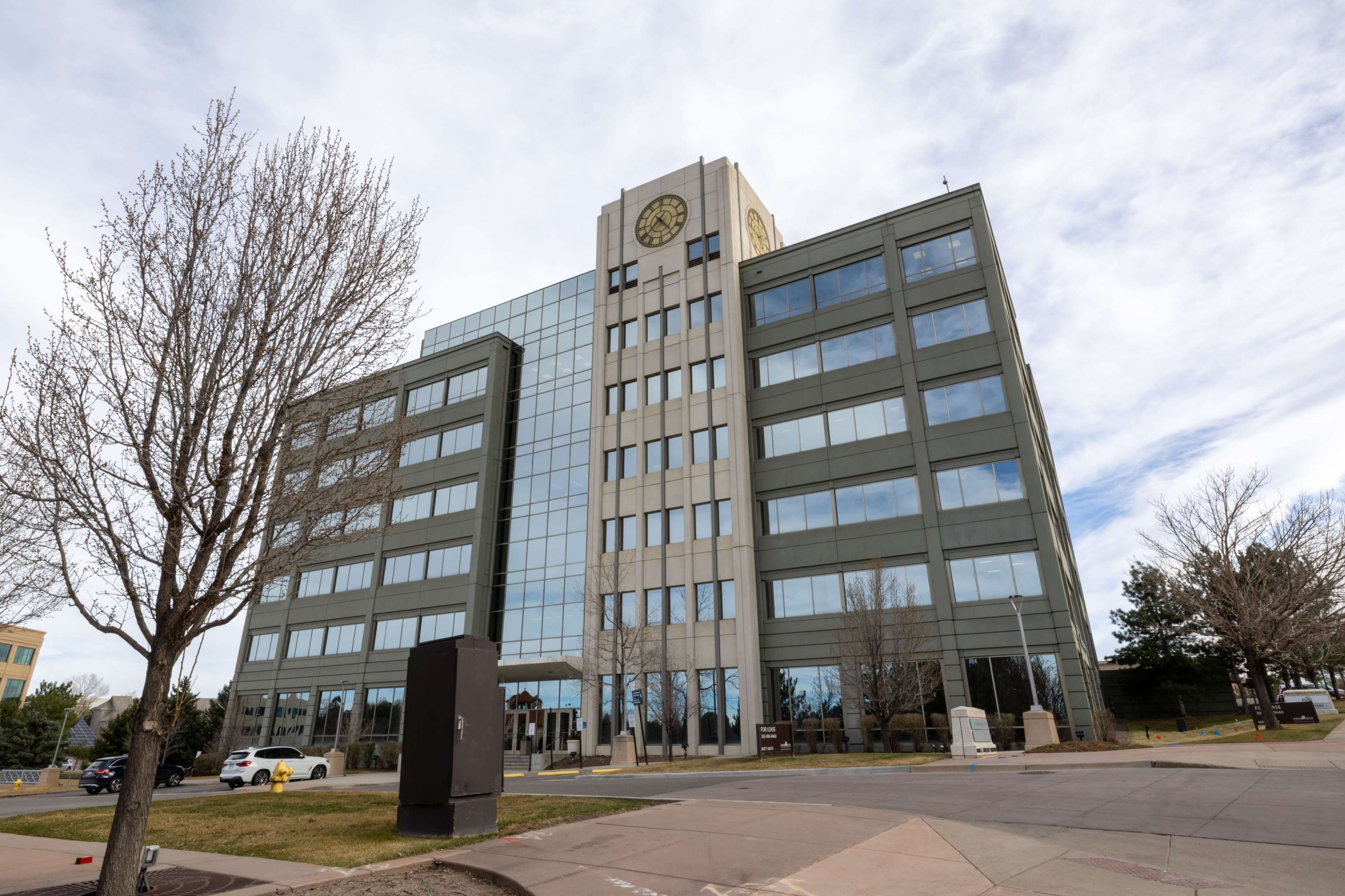 A modern office building with a large clock is situated in an open area, featuring large glass windows and a landscaped entrance.