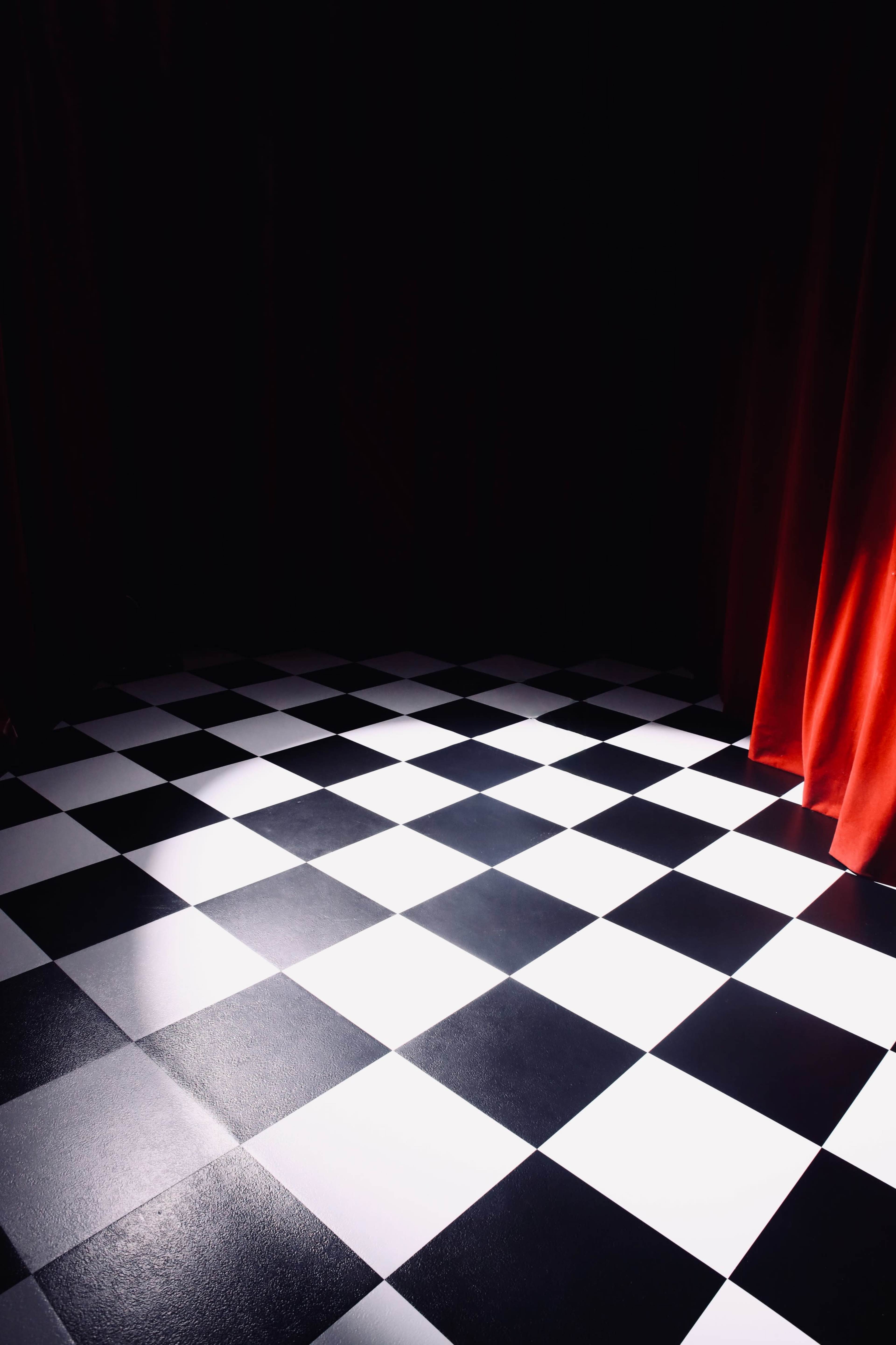 The image shows a checkerboard-patterned floor with alternating black and white squares, illuminated against a dark backdrop and partially draped by a red curtain.