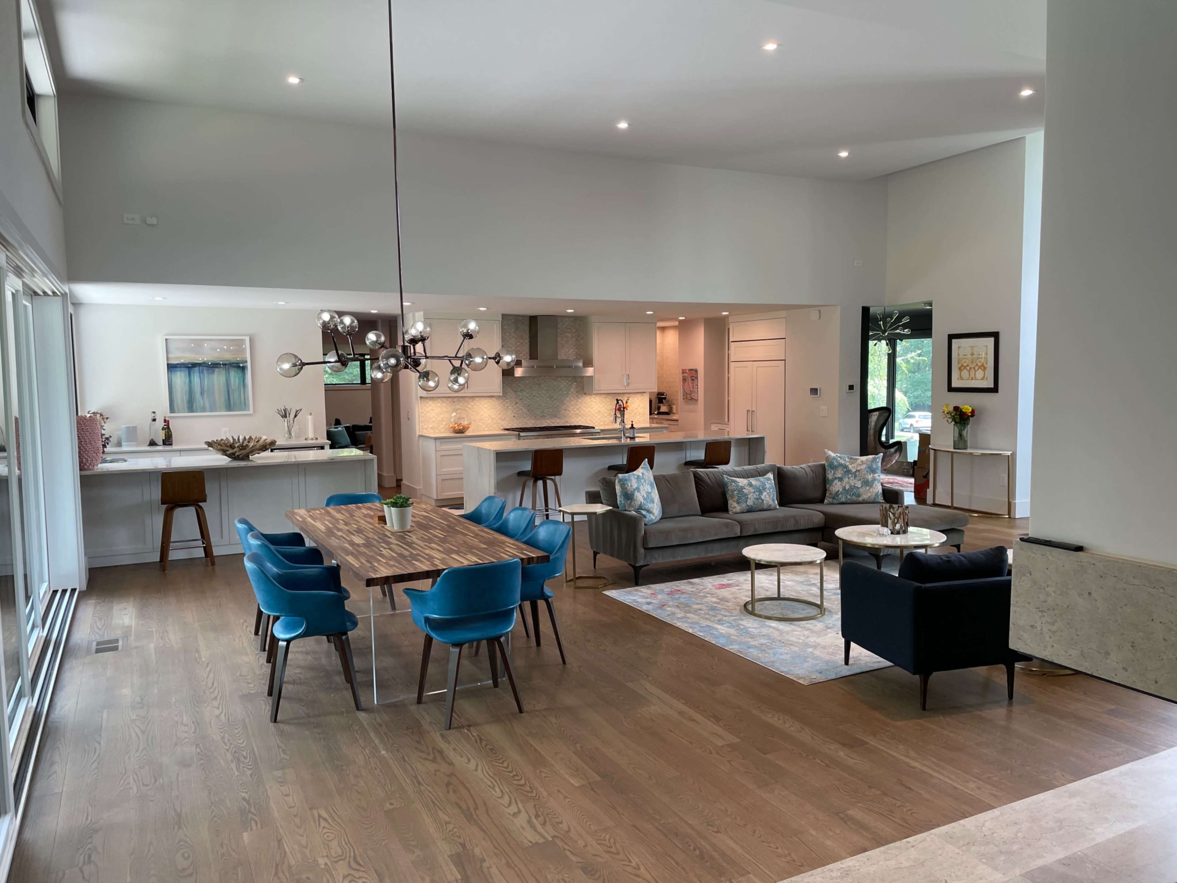 The image shows a spacious living area with a dining table, blue chairs, and a modern kitchen in the background.