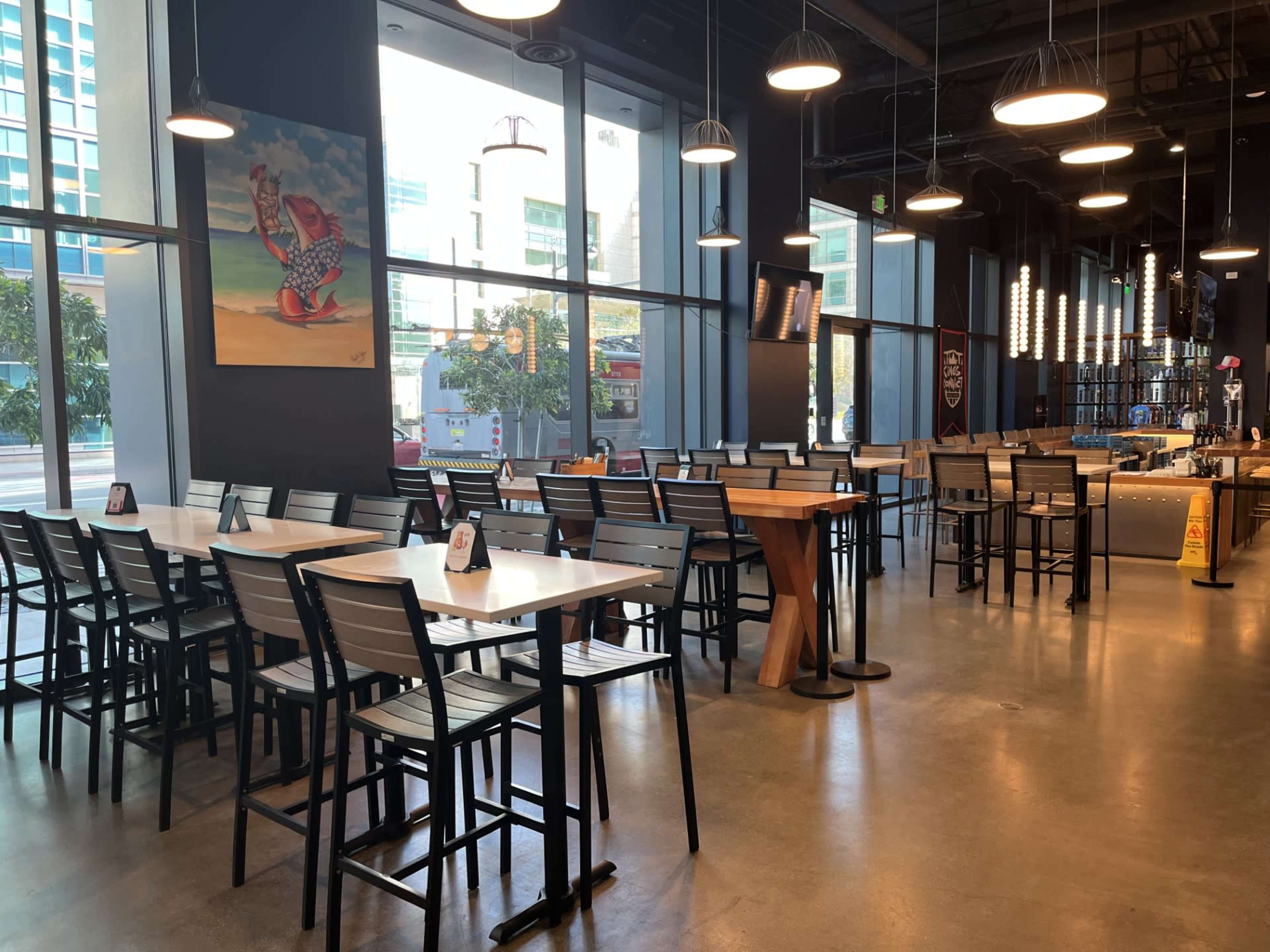 The image shows a modern restaurant interior with multiple tables and chairs, large windows, and a bar area in the background.