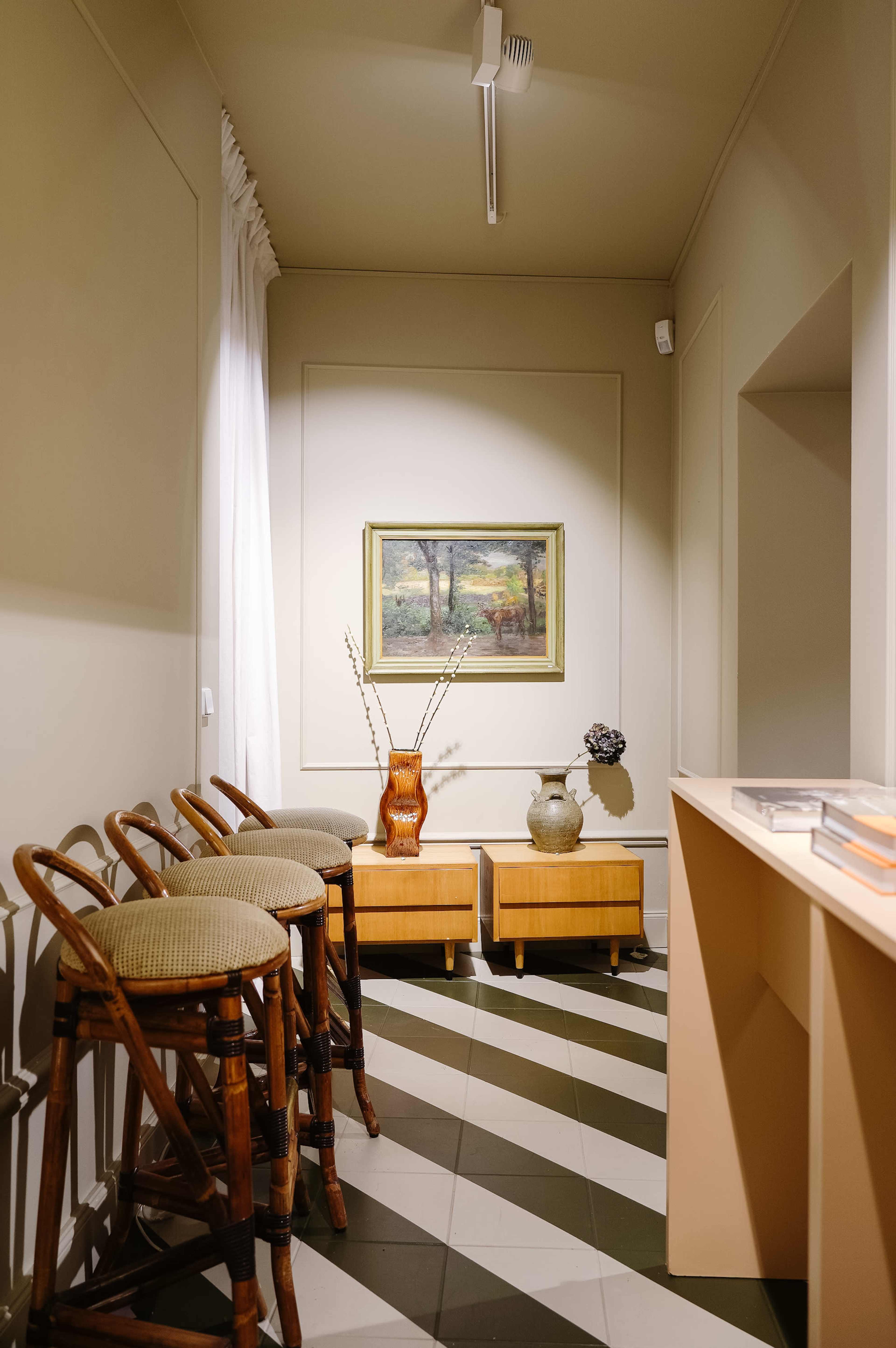 A minimalist interior features three wooden bar stools, a painting on the wall, and a console table with a vase and a flower arrangement.