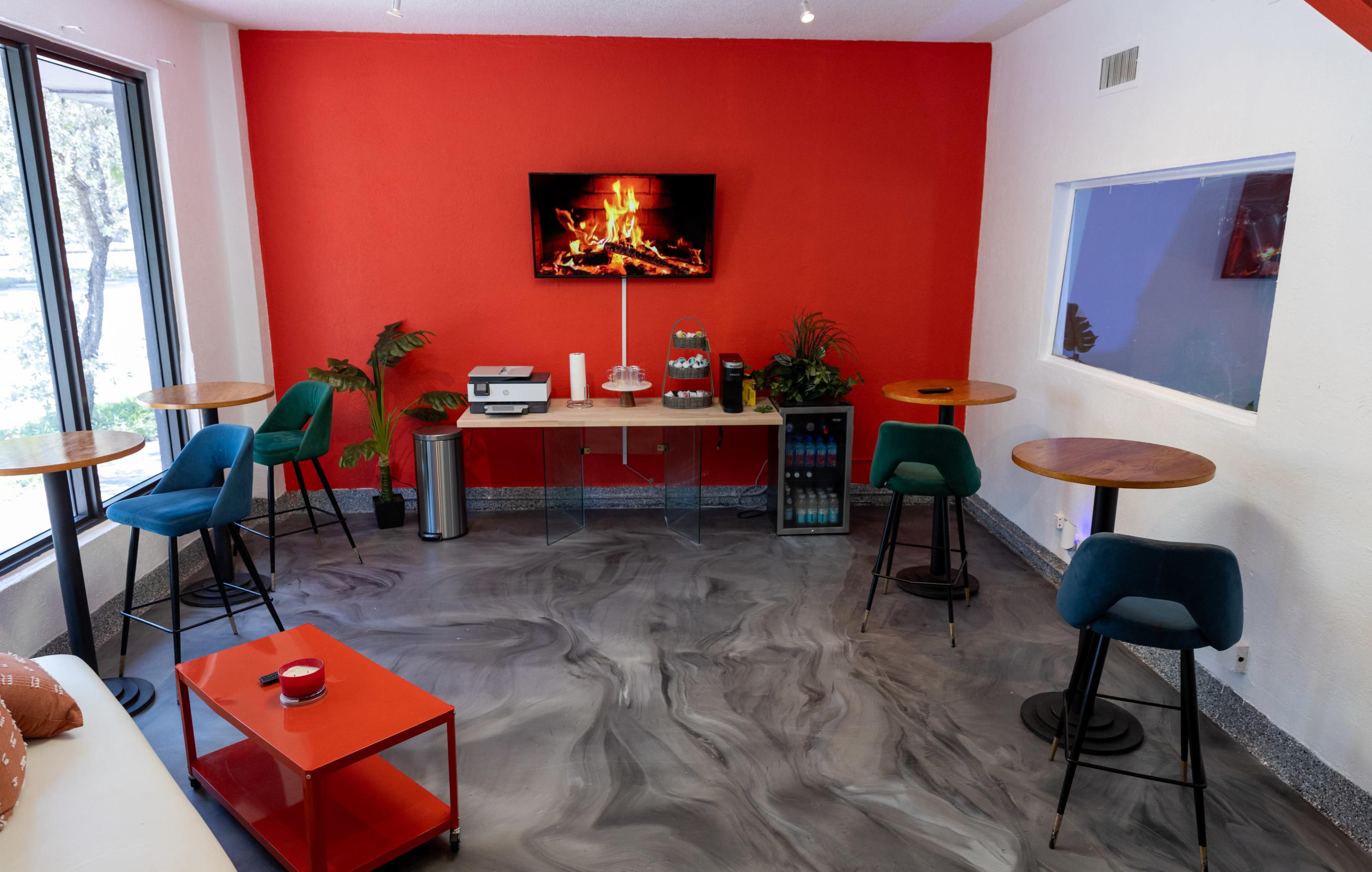 A cozy lounge area features a television displaying a fireplace, surrounded by small tables and chairs against a bright red wall.