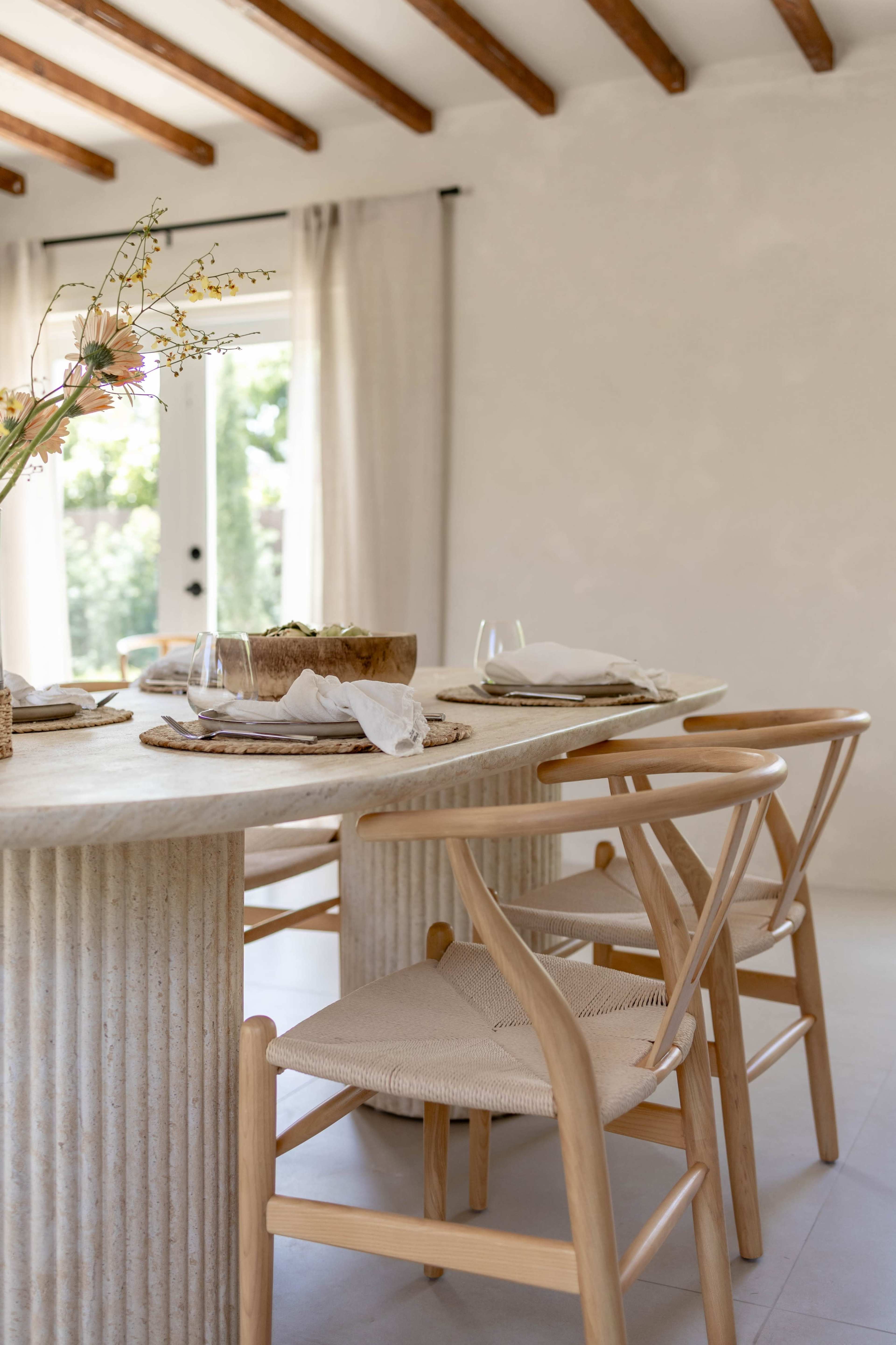 A modern dining table with a textured base is set with plates and glasses, accompanied by natural decor and a view of a sunny outdoor space through large windows.