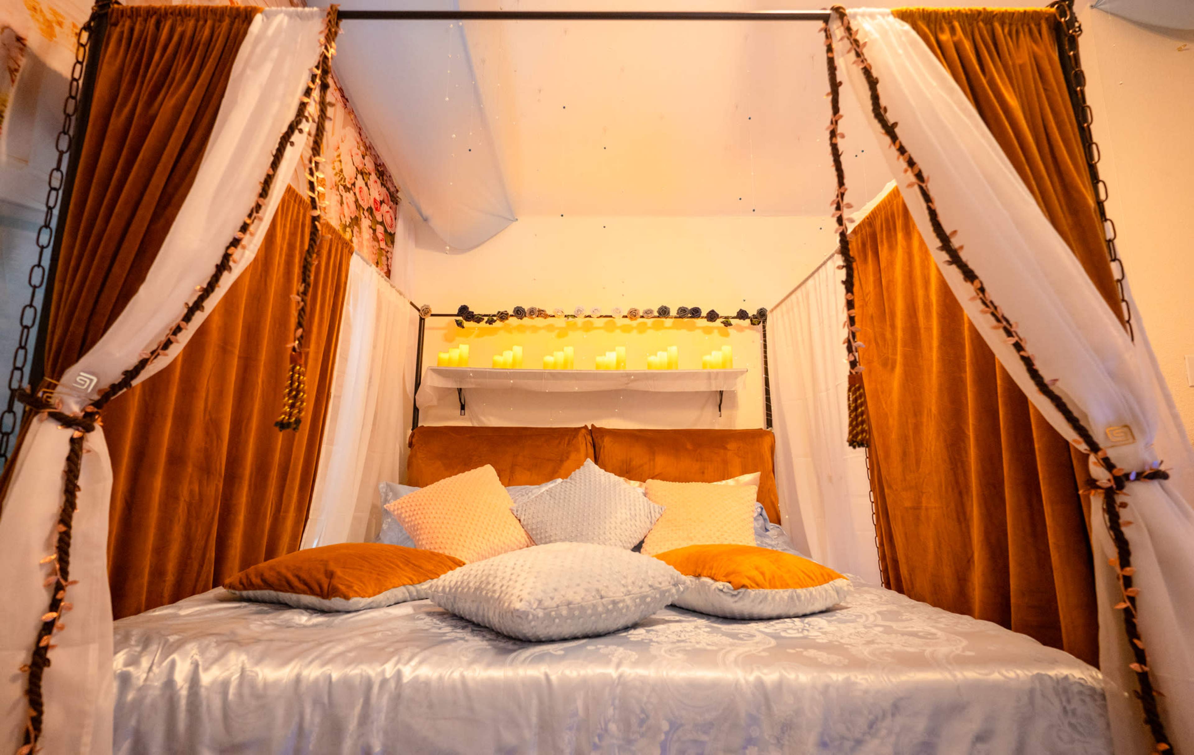 The image shows a cozy bed with a wooden frame, draped with soft curtains, and adorned with various pillows in a warm-toned setting.