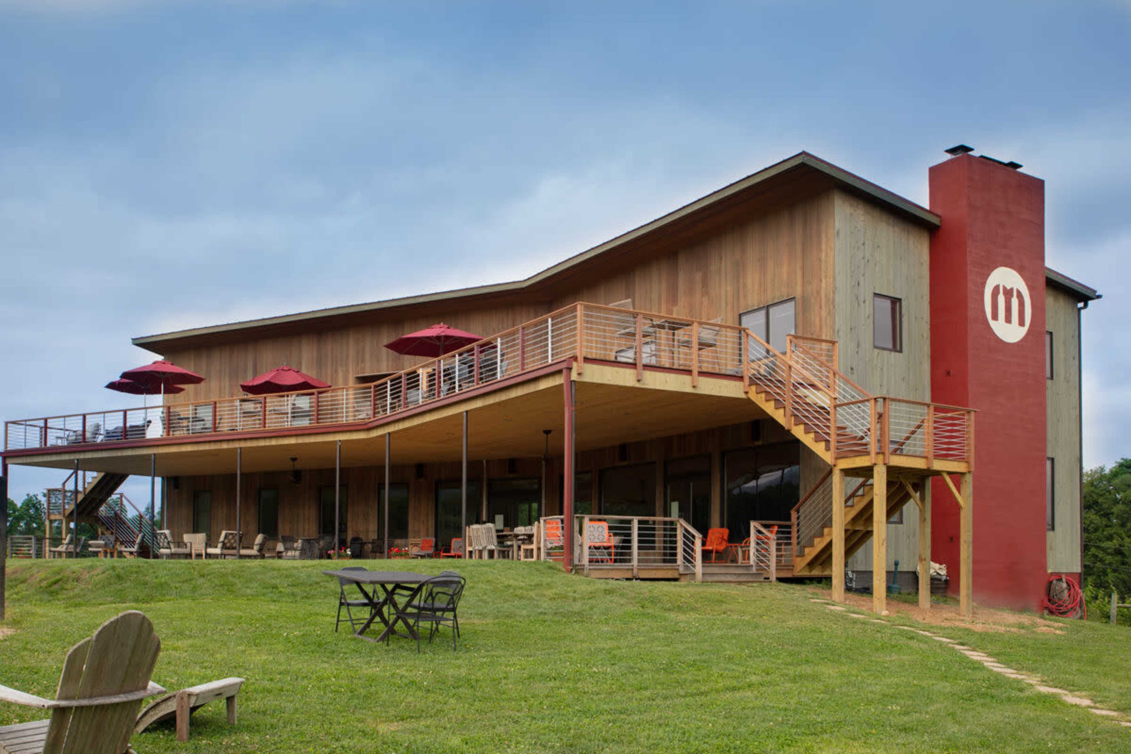 The image shows a modern wooden building with a red accent wall and a wraparound deck featuring outdoor seating and umbrellas.