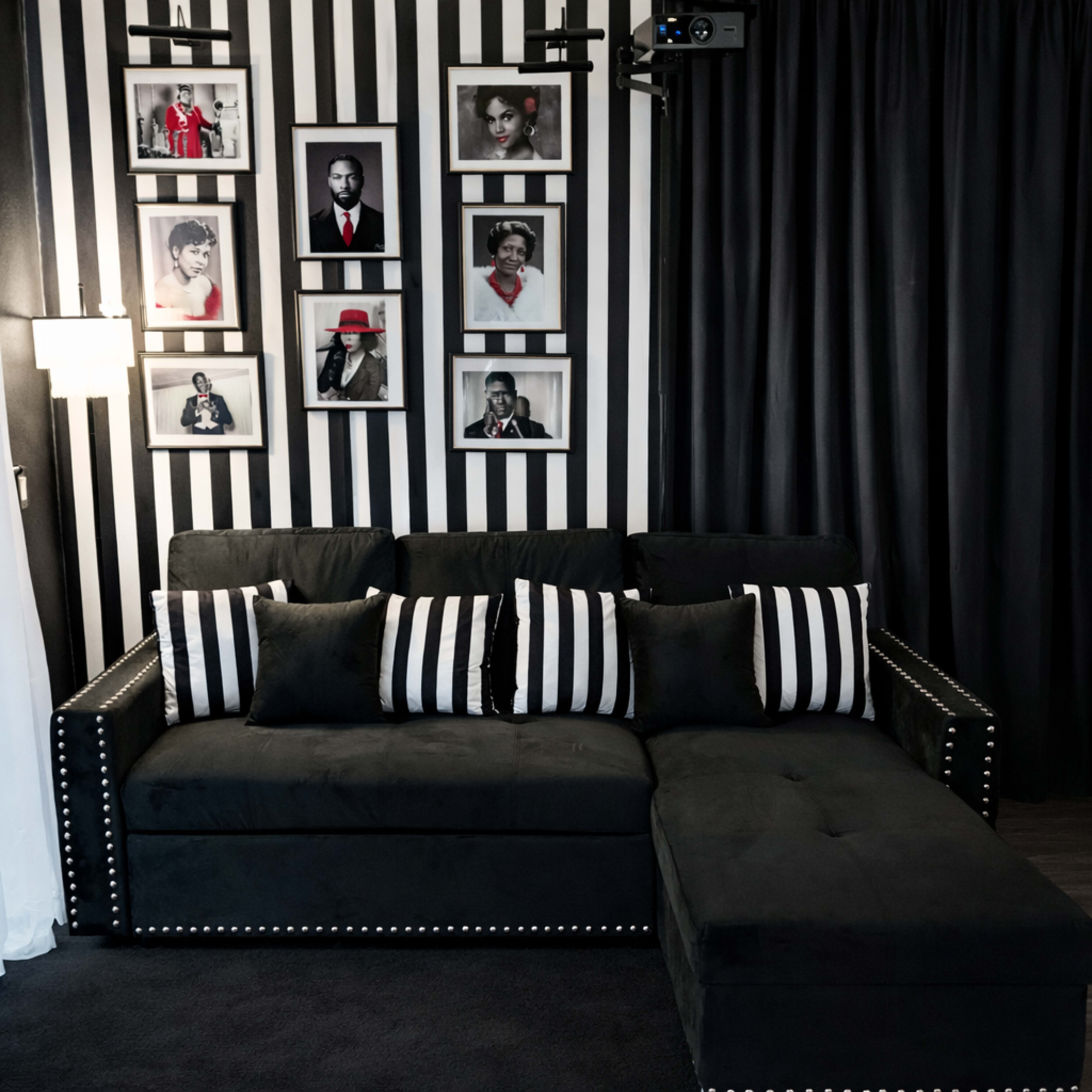 The image shows a black sectional sofa with striped throw pillows, positioned against a wall featuring black and white striped wallpaper and framed black-and-white portraits.