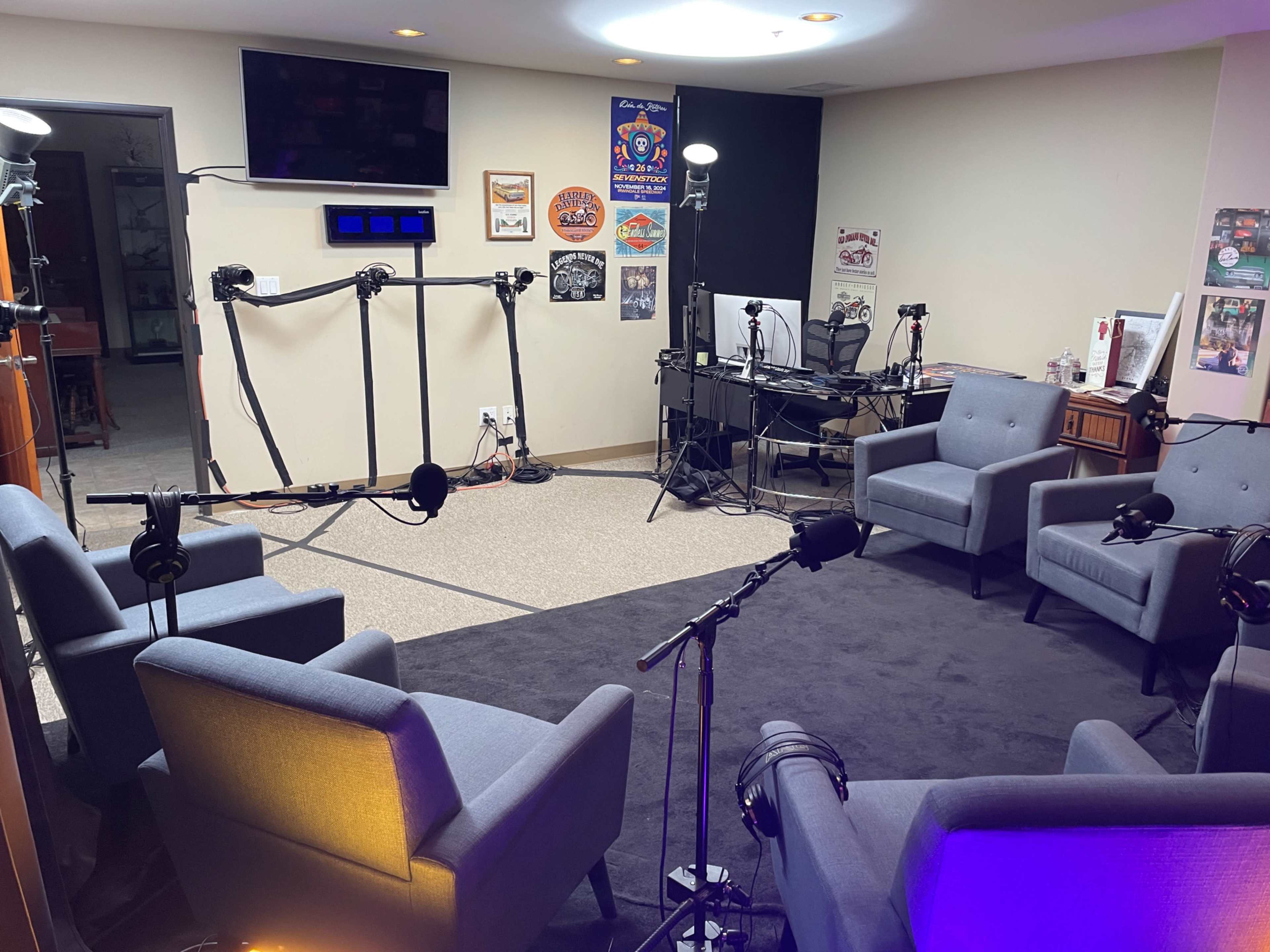 The image shows a recording studio setup with four gray chairs arranged in a circular layout, surrounded by microphones on stands and a desk with equipment in the background.