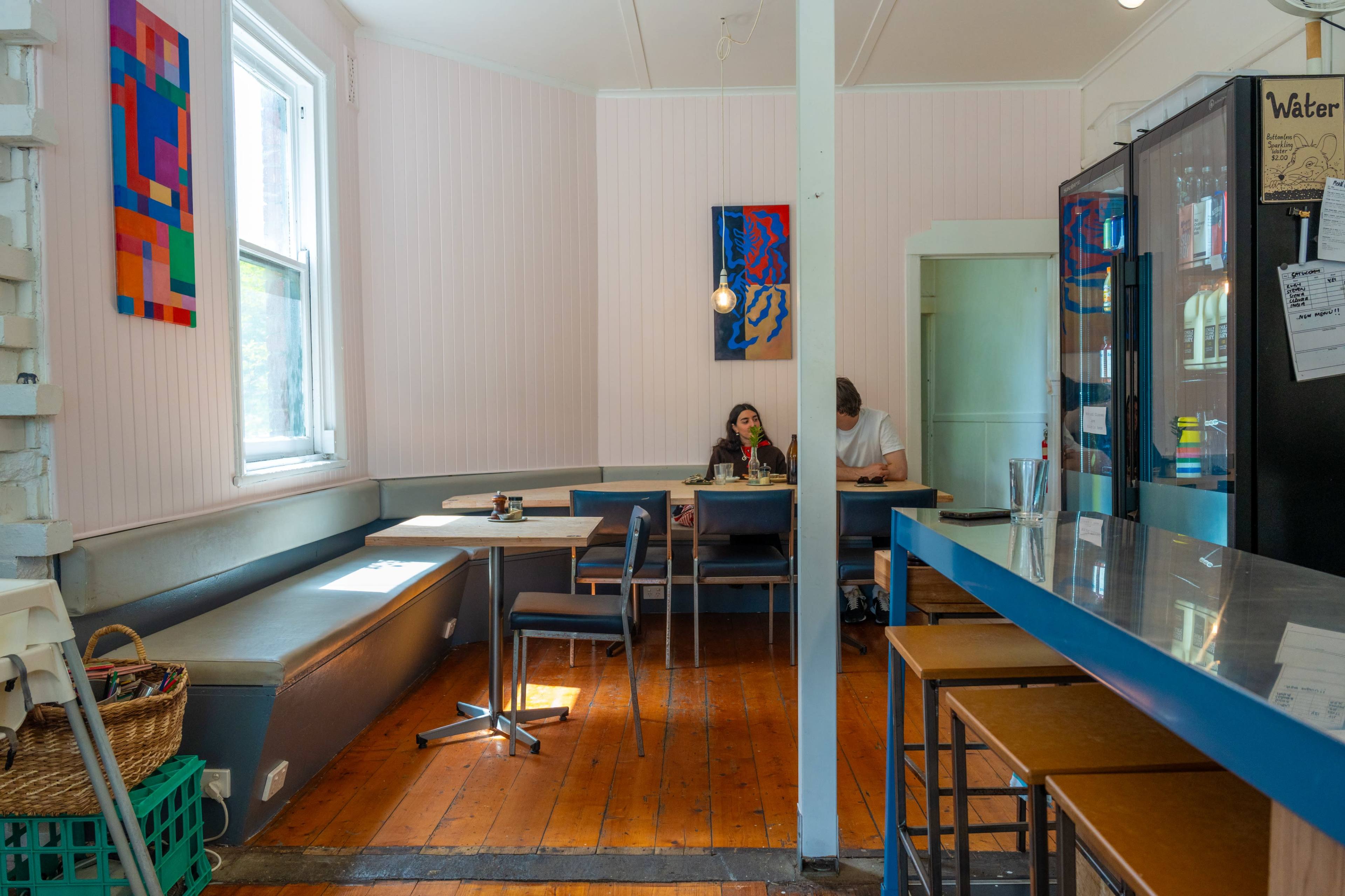 The image shows a cozy café interior with a mix of seating arrangements, including a booth and table, and features colorful artwork on the walls.