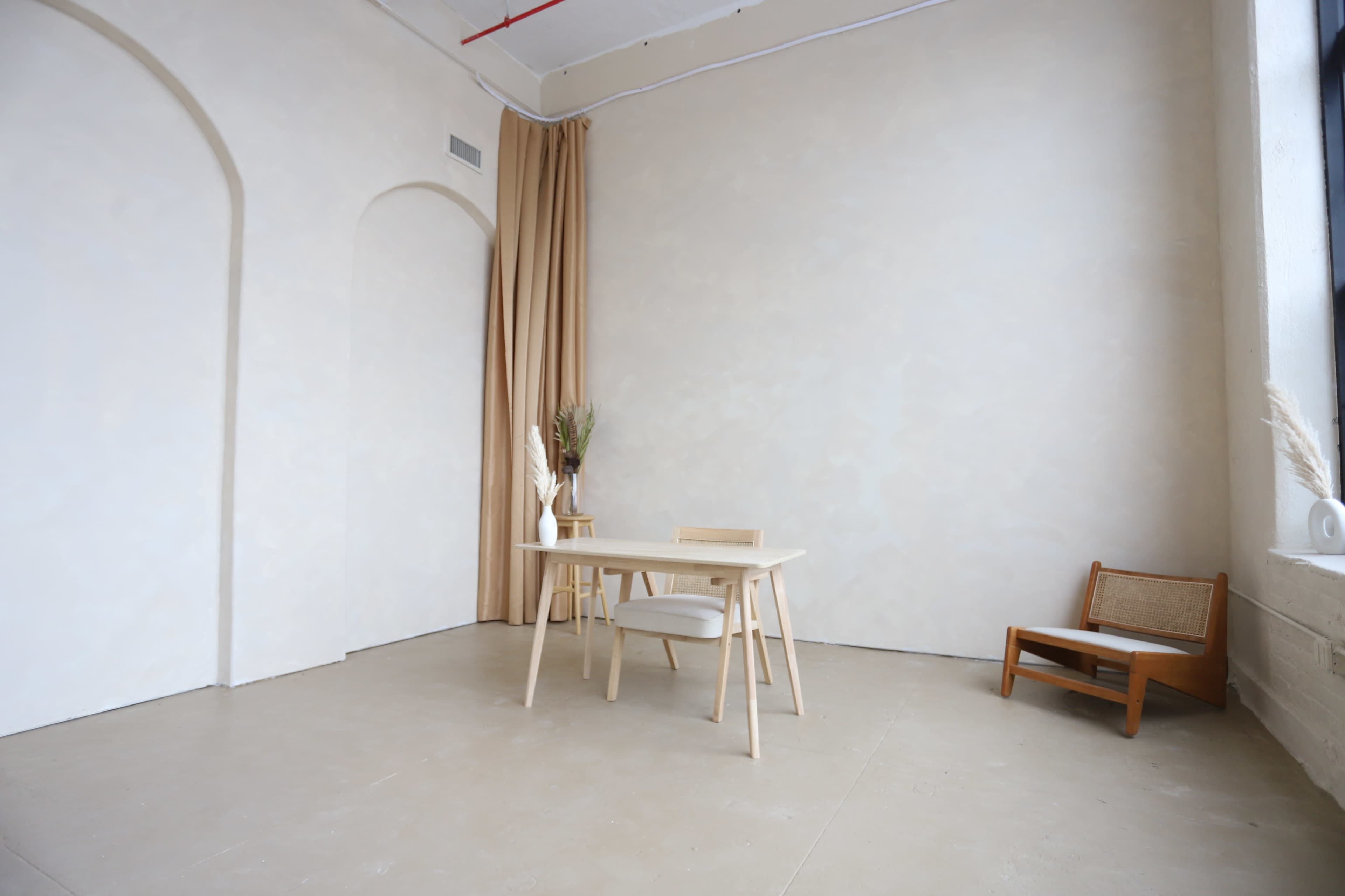 A minimalistic room features a light-colored wall, a wooden table with two chairs, a decorative plant, and a small chair in the corner.