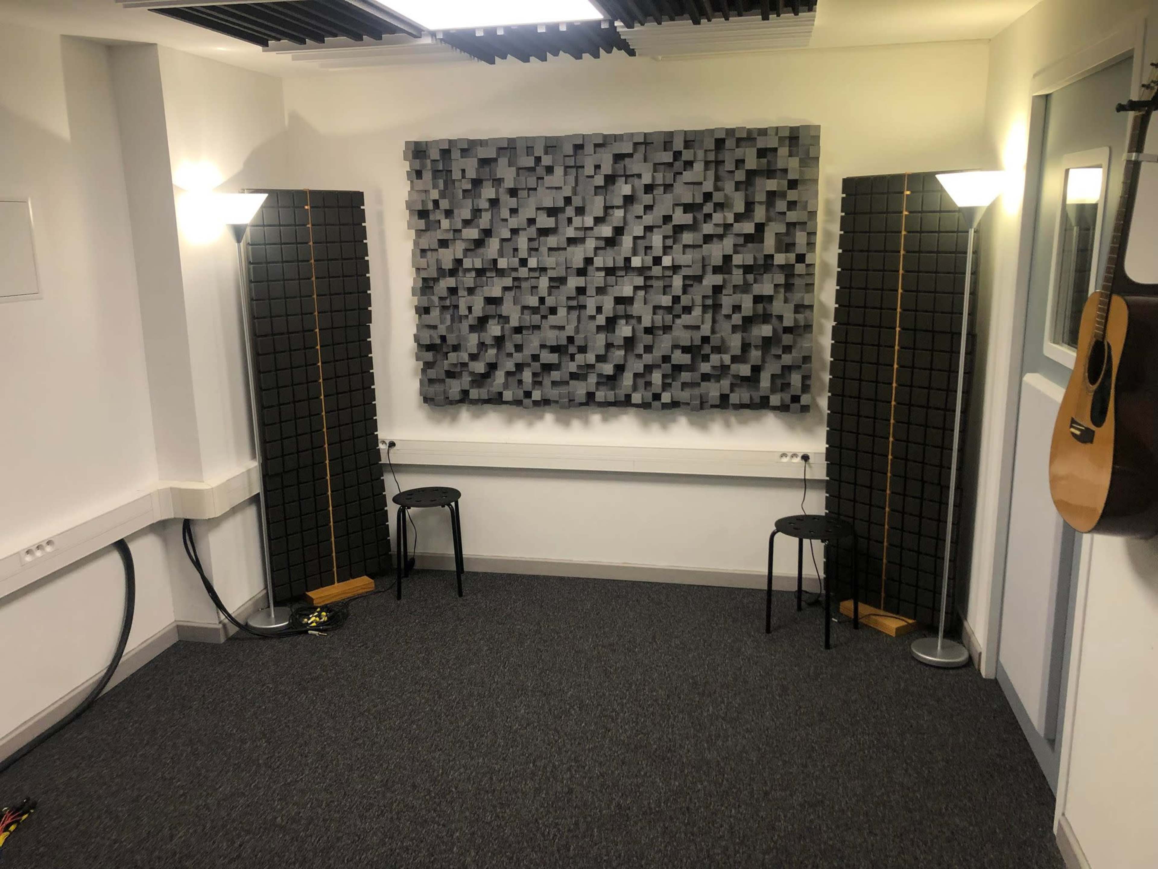 A small soundproof room featuring two black acoustic panels, two black stools, and a guitar hanging on the wall.
