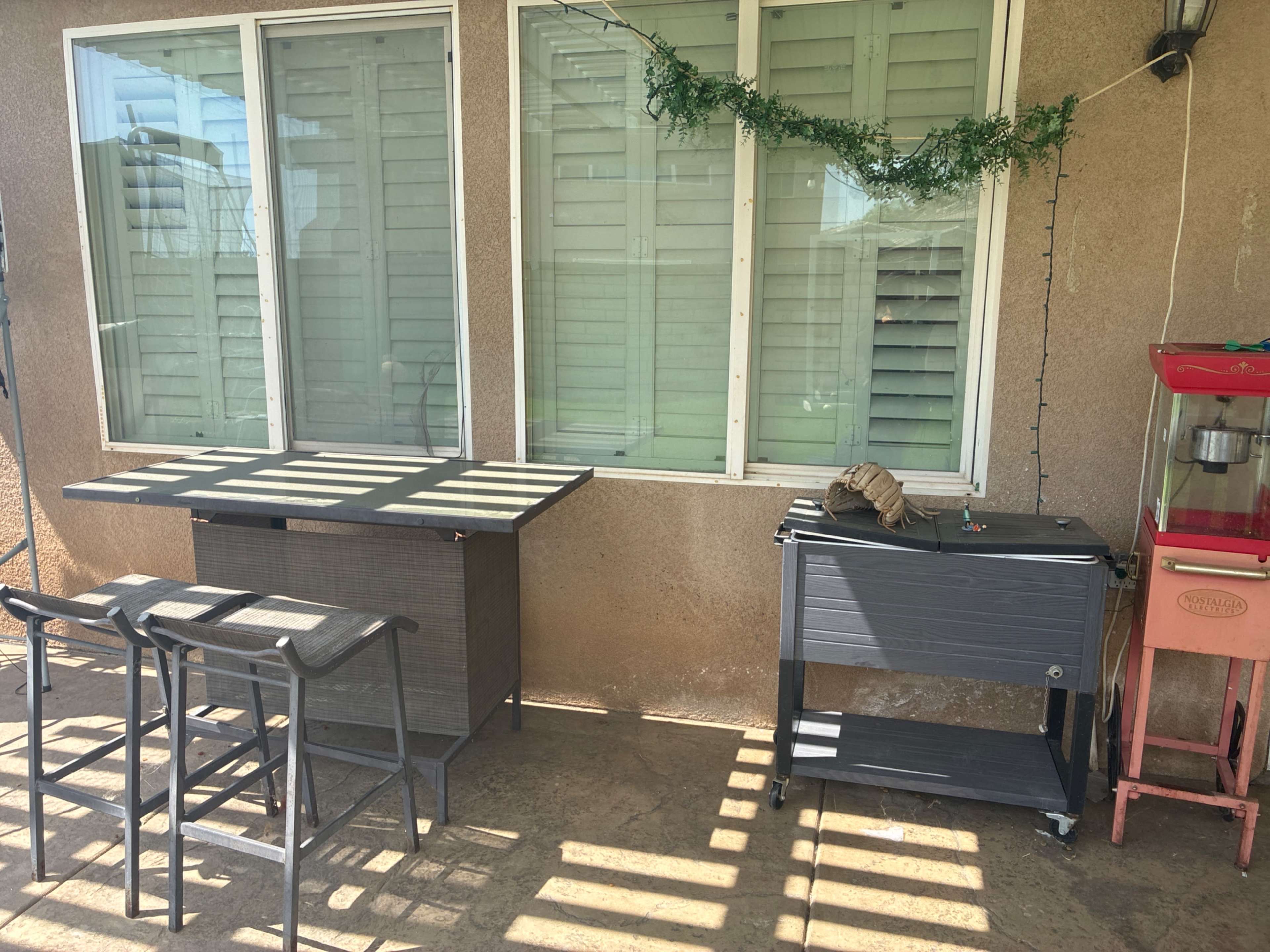 The image shows a patio area with a table and two bar stools on one side, and a small cart next to a popcorn machine against the wall, all partially shaded by diagonal sunlight.