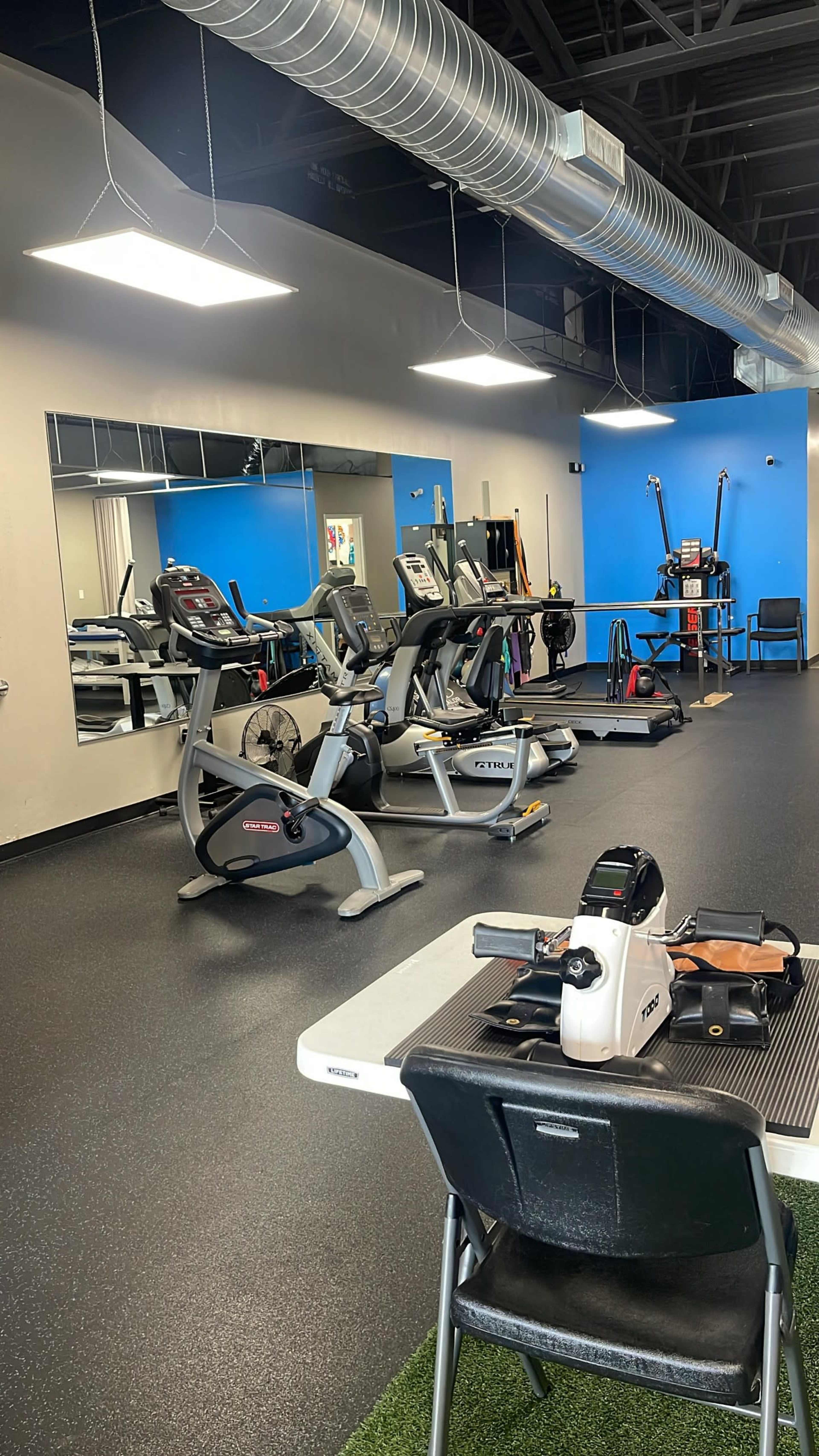 The image shows a gym interior featuring exercise equipment, a mirrored wall, and a table with a chair in the foreground.
