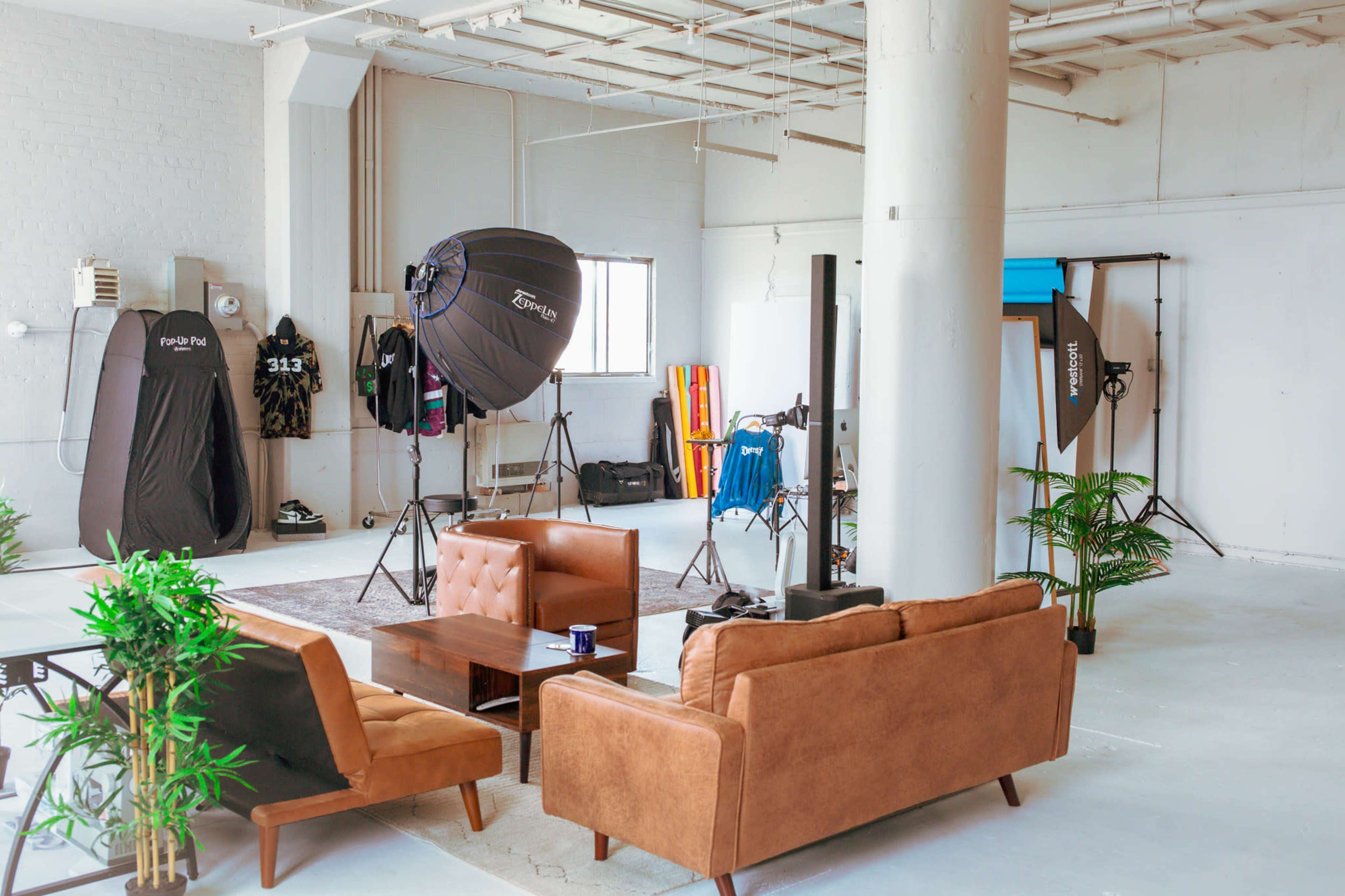 A spacious studio with a brown couch, a coffee table, and photography equipment set up around the room, including softbox lights and backdrops.