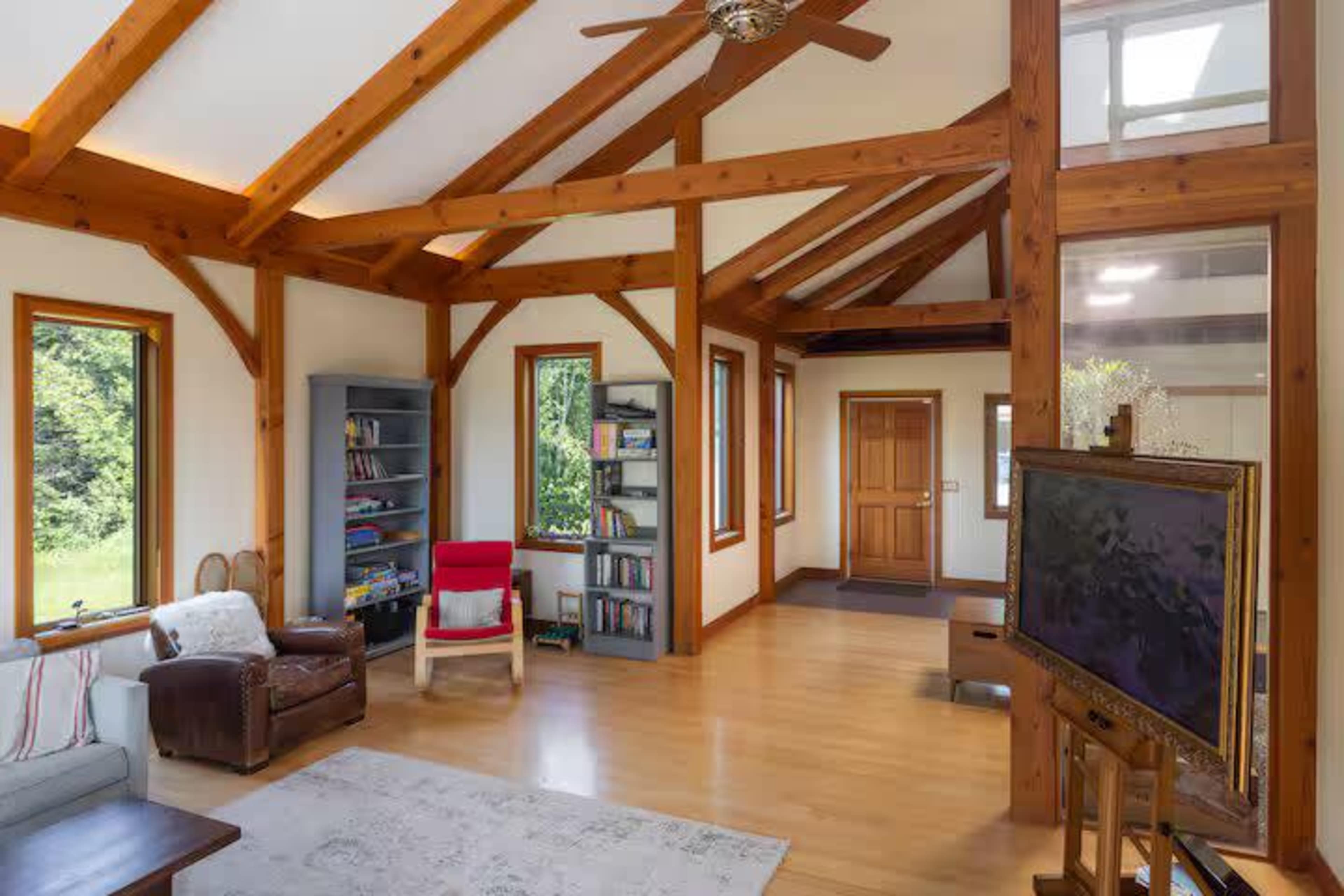The image shows a spacious, modern living room with wooden beams, a comfortable seating area, a bookcase, and a large window allowing natural light.
