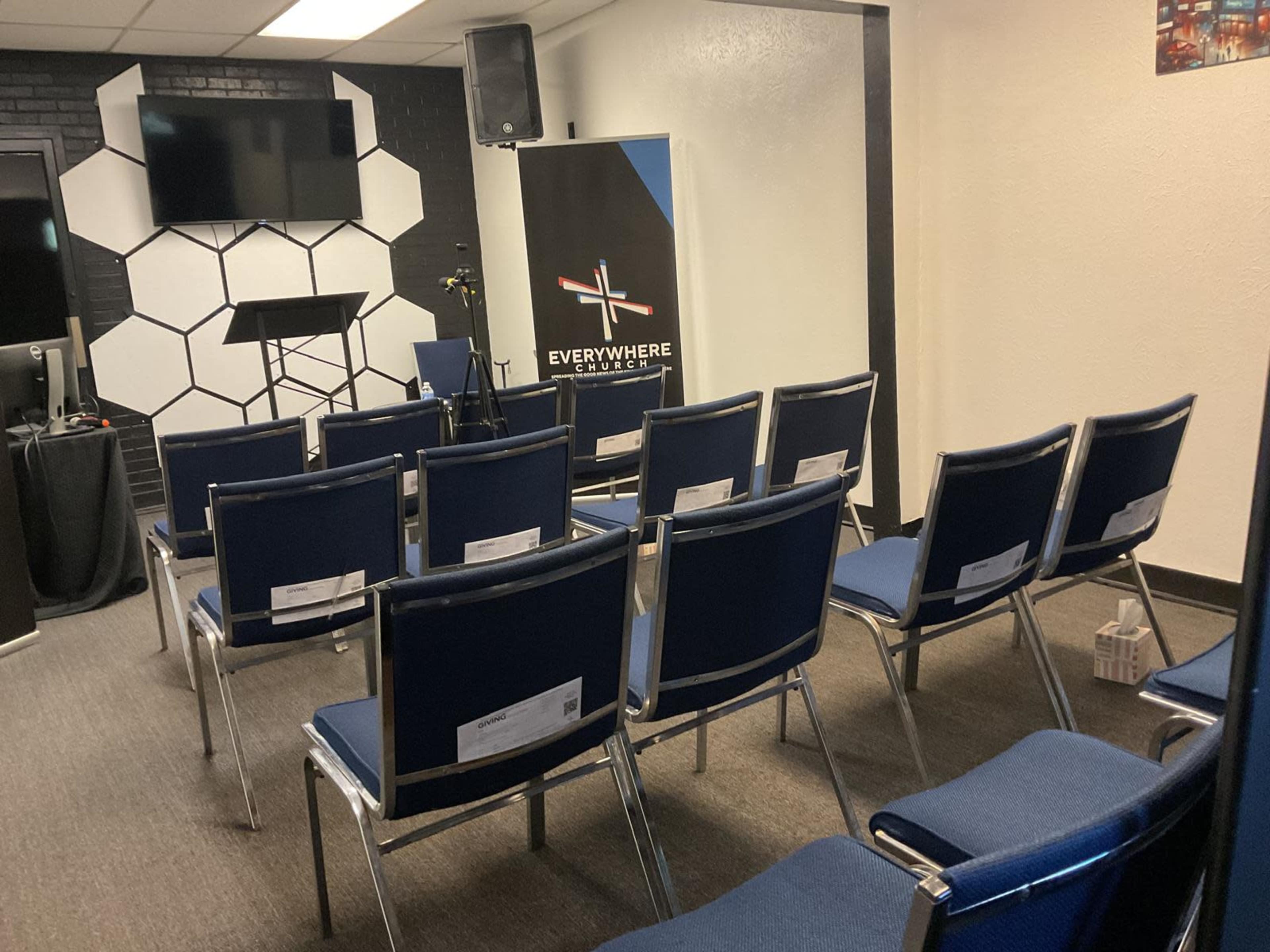 The image shows a room set up with metal-framed chairs arranged in rows, facing a blank projection screen and a banner advertising an organization called "Everywhere."