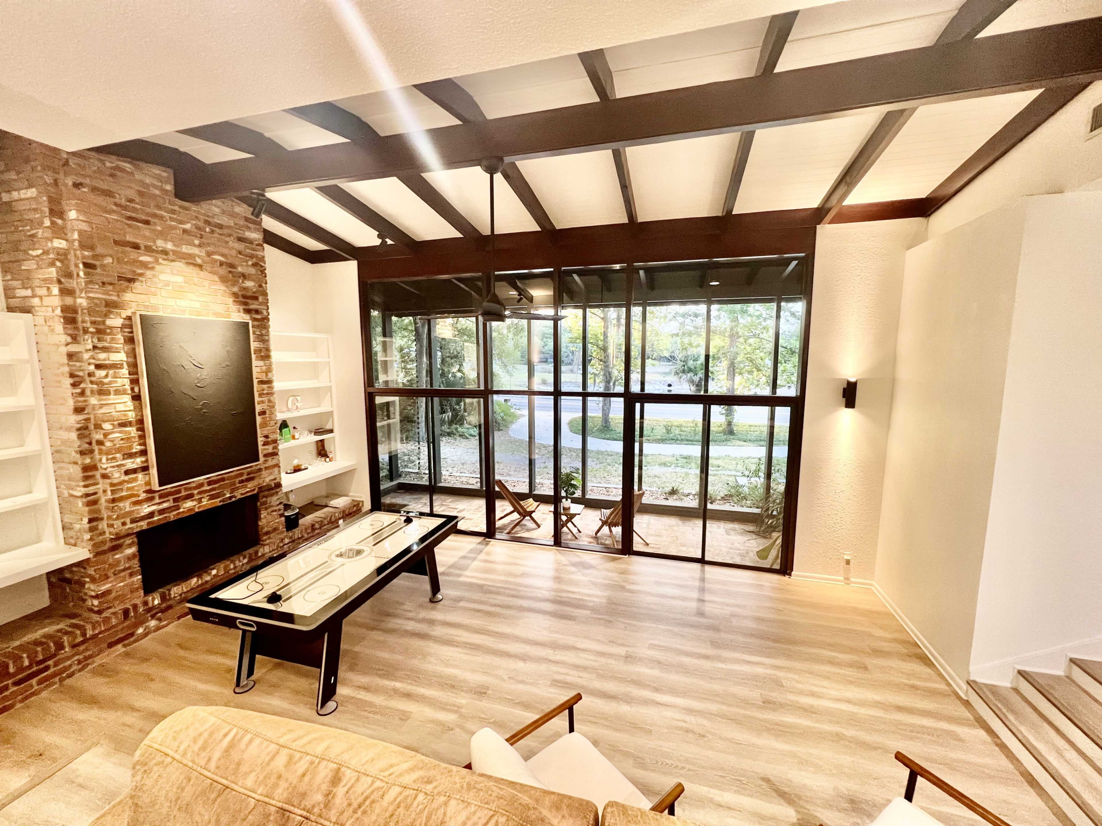 A modern living room with a large glass wall overlooking an outdoor garden area and featuring a brick accent wall, wooden beams, and a coffee table in the center.