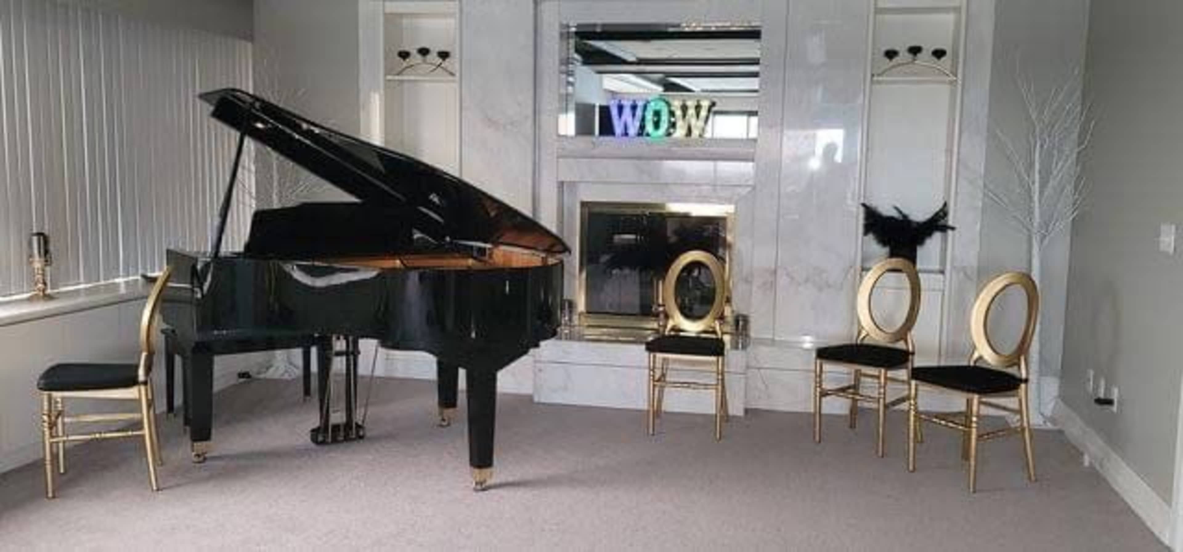 A black grand piano is positioned next to a fireplace in a minimalist room, accompanied by three gold and black chairs.