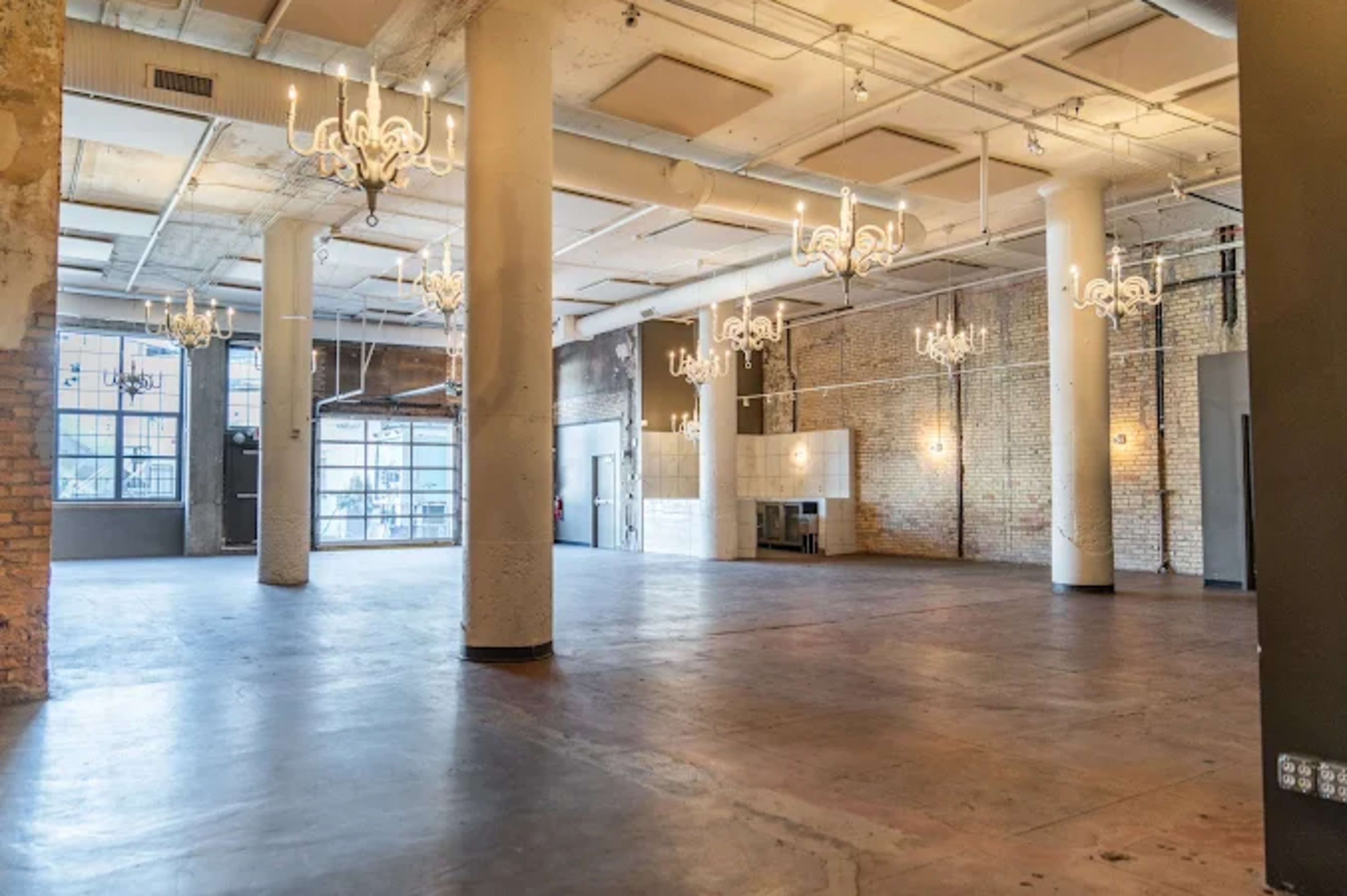 The image shows a spacious, industrial-style room with polished concrete floors, exposed brick walls, and several chandeliers hanging from the ceiling.