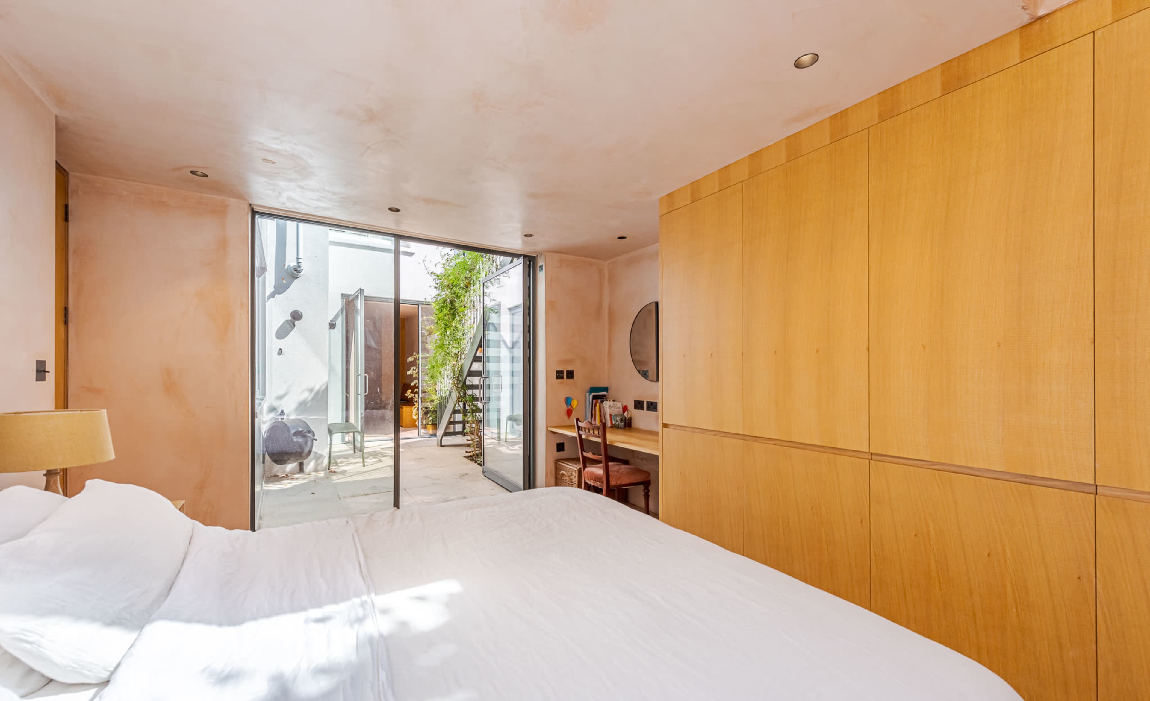 A minimalist bedroom with a large bed and a wooden wall unit, featuring a glass door that opens to a patio area.