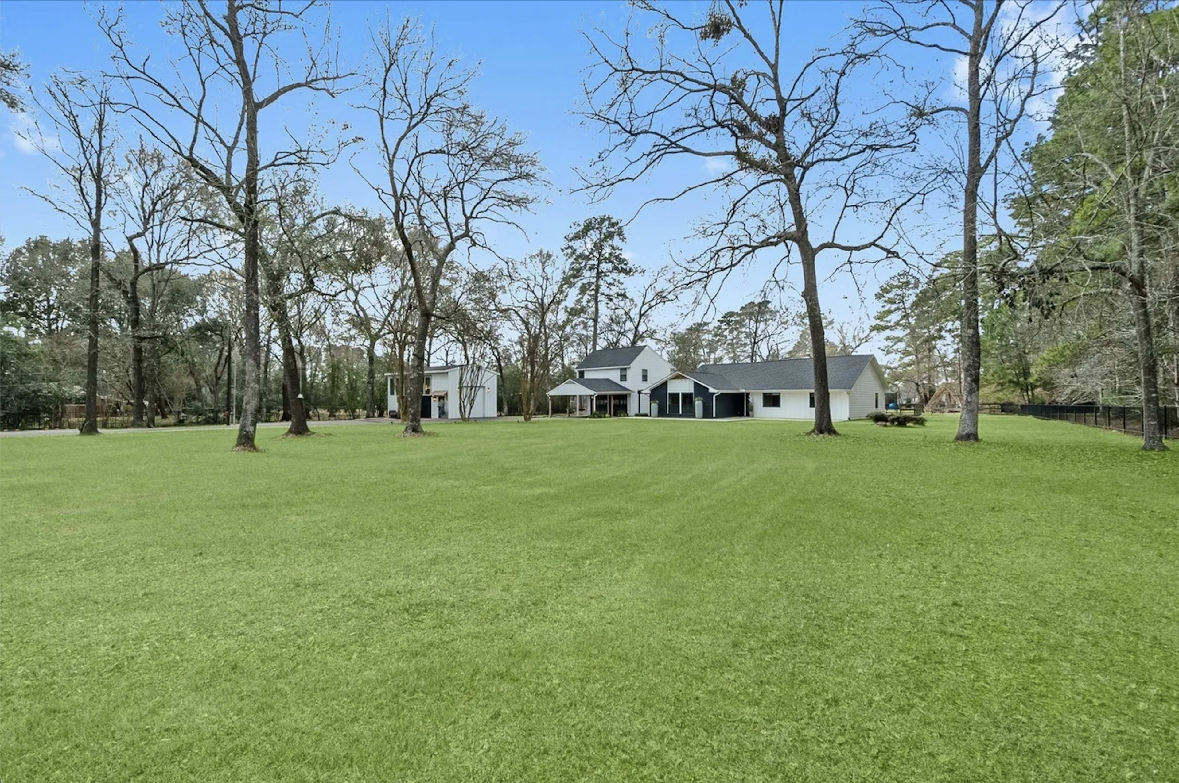 A spacious grassy area is surrounded by trees, with a white house and a black-roofed house in the background.