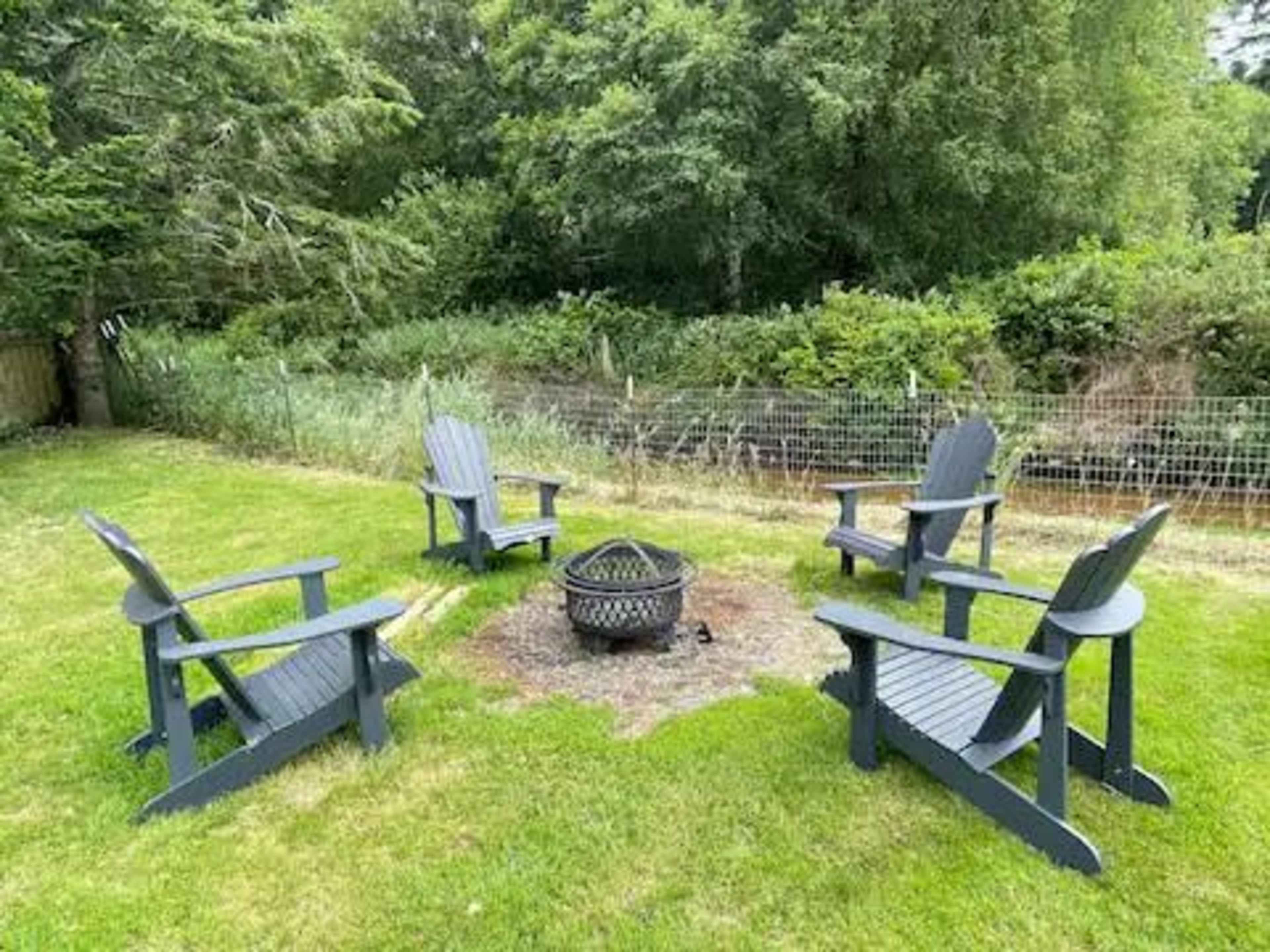 The image features a fire pit surrounded by four black Adirondack chairs on a grassy area, with greenery and a fence in the background.