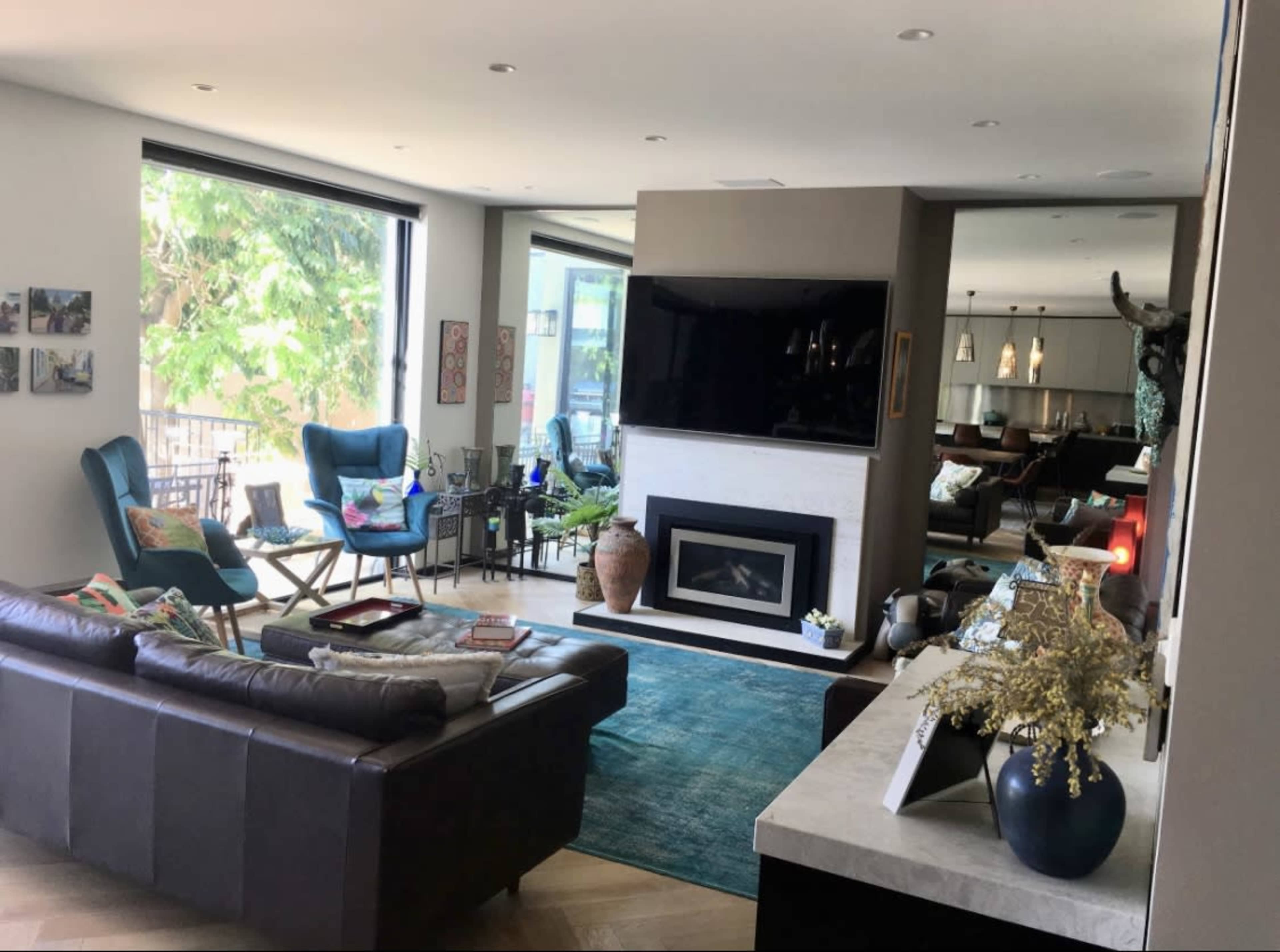 A modern living room with a large TV above a fireplace, blue chairs, and a coffee table, featuring natural light from large windows.