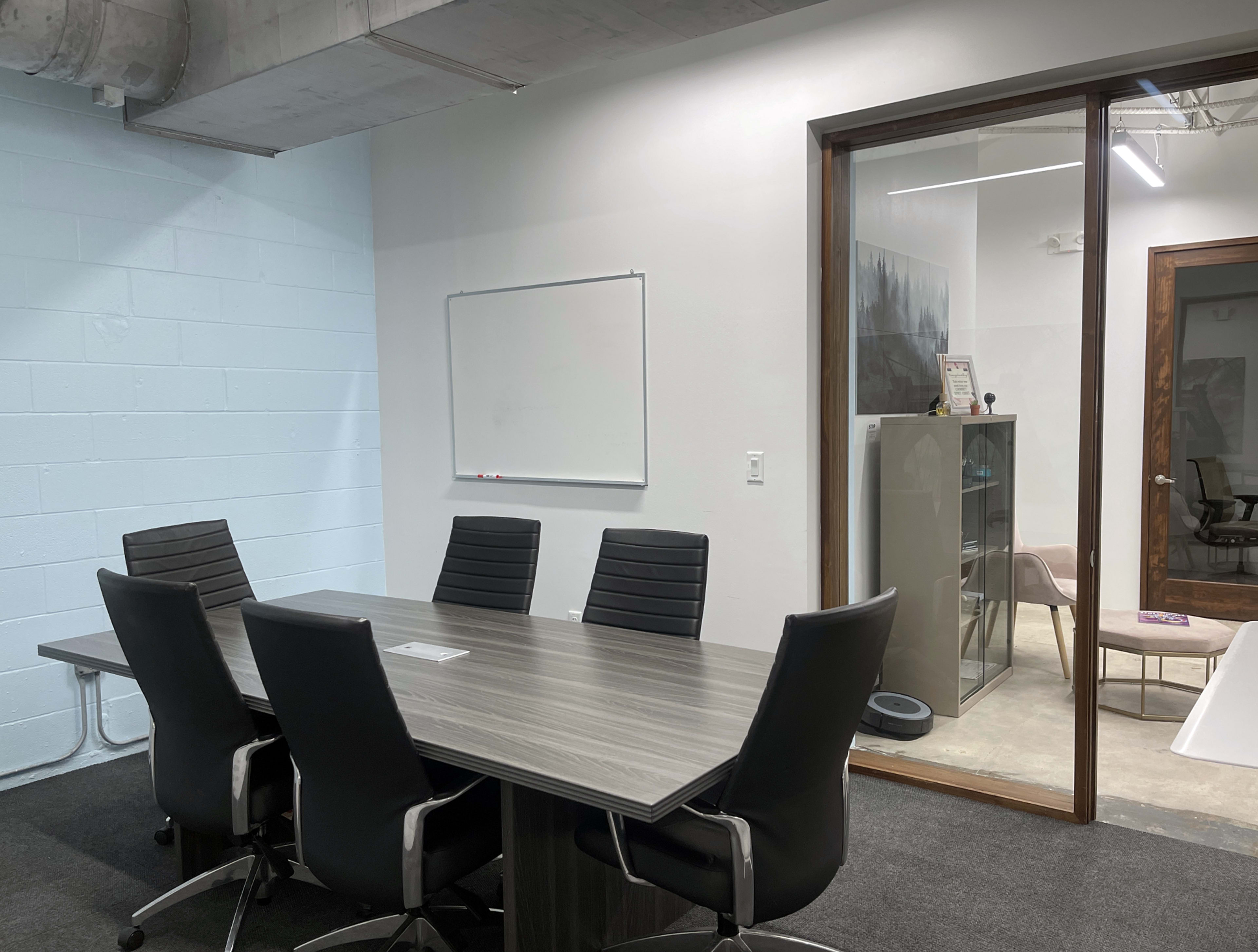 The image shows a conference room with a rectangular table and six black chairs, complemented by a whiteboard on one wall and a glass-walled office in the background.