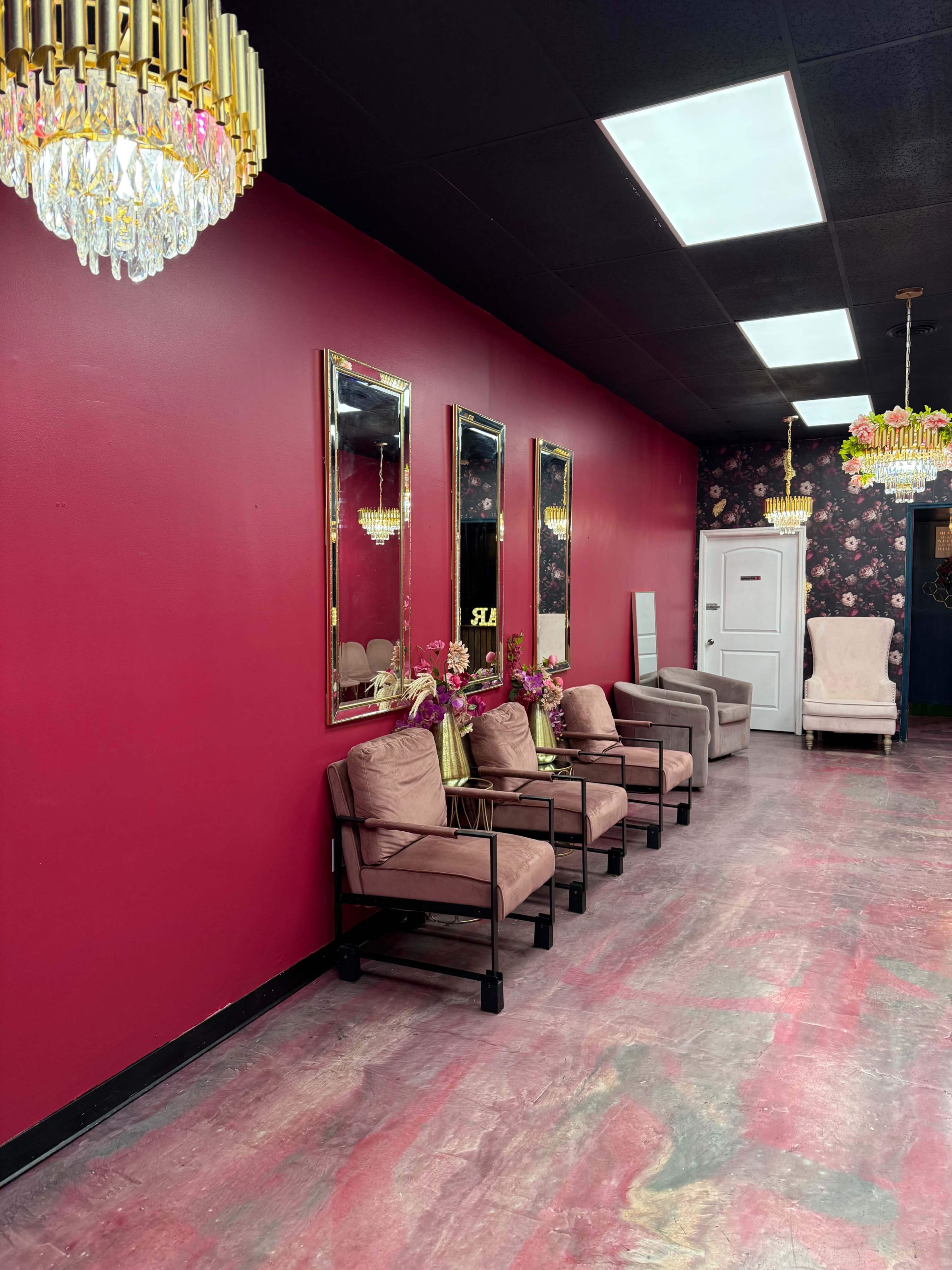 The image shows a salon interior with a maroon wall, three mirrors, elegant light fixtures, and a row of beige seating along the floor.