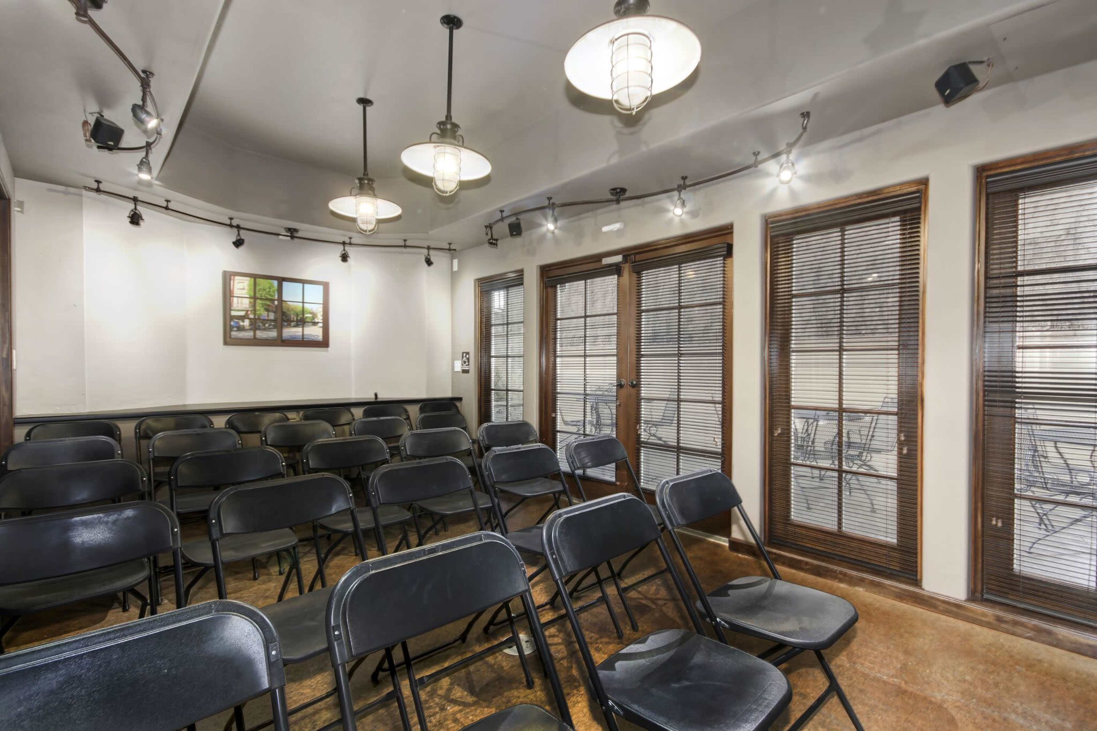 A meeting room arranged with black folding chairs facing a set of windows with blinds and a framed picture on the wall.