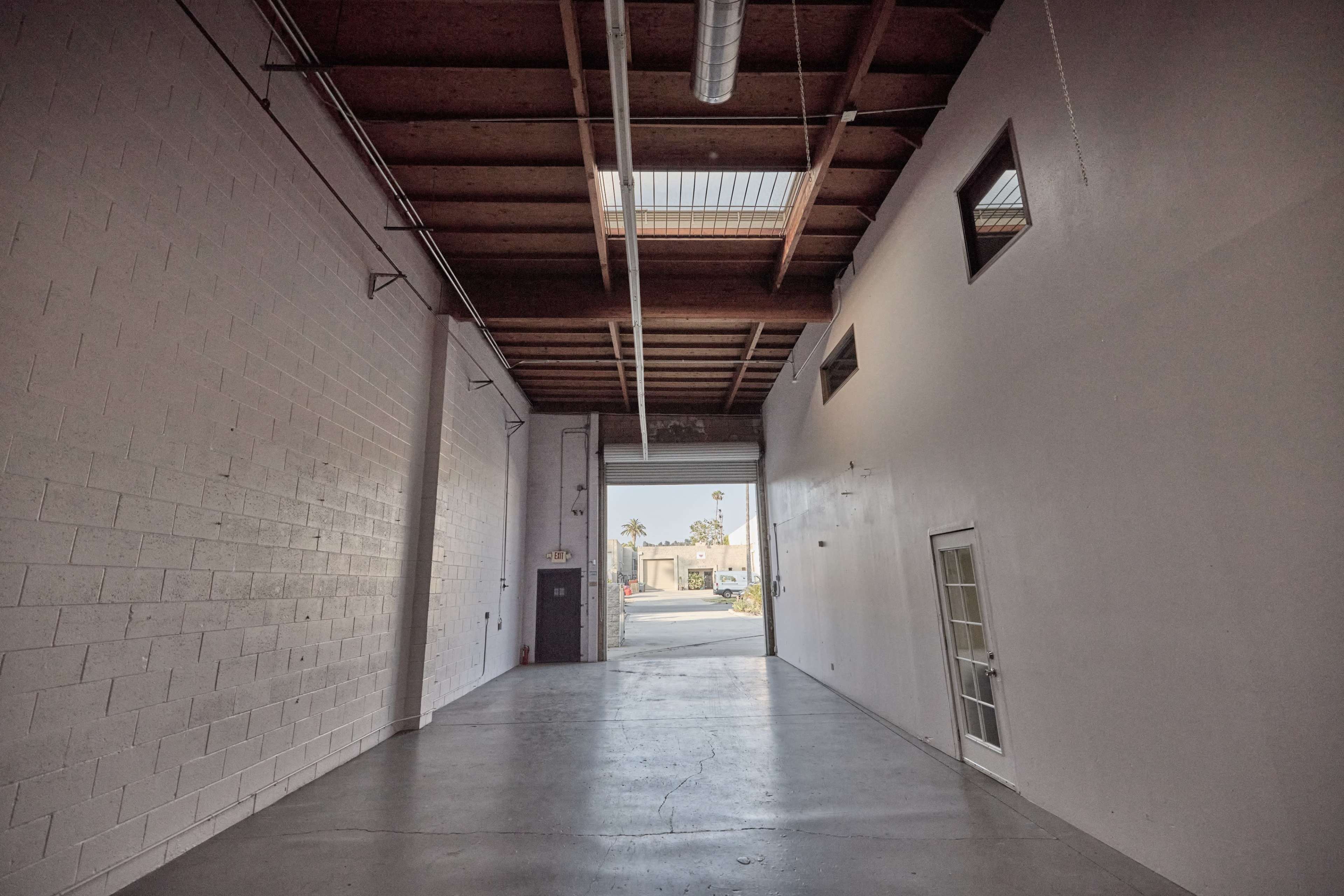 A wide, empty corridor extends from a brightly lit entrance to a plain white wall, with a door and small windows on either side.