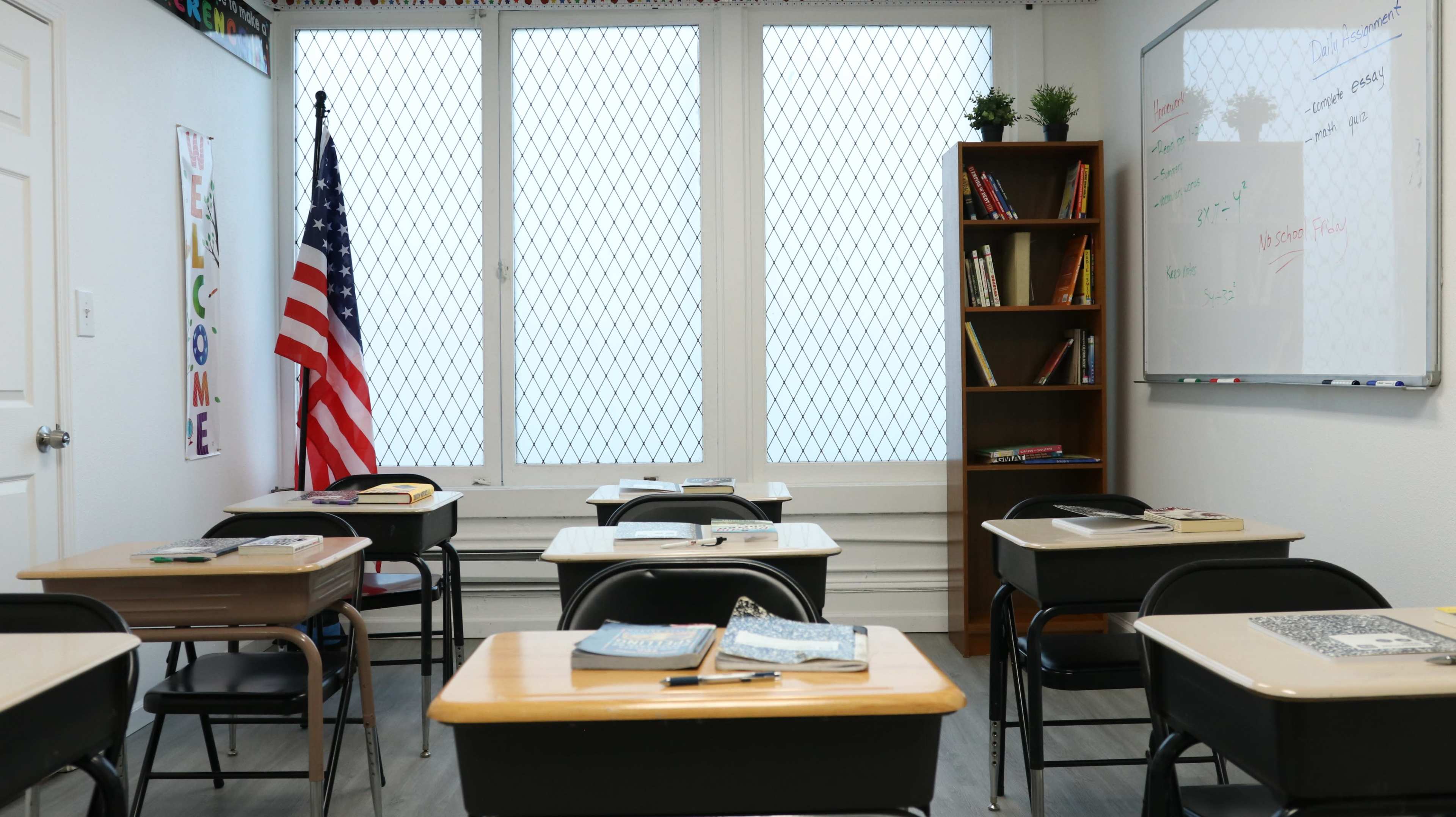 A classroom is set up with several desks, an American flag, a bookcase filled with books, and a whiteboard on the wall.