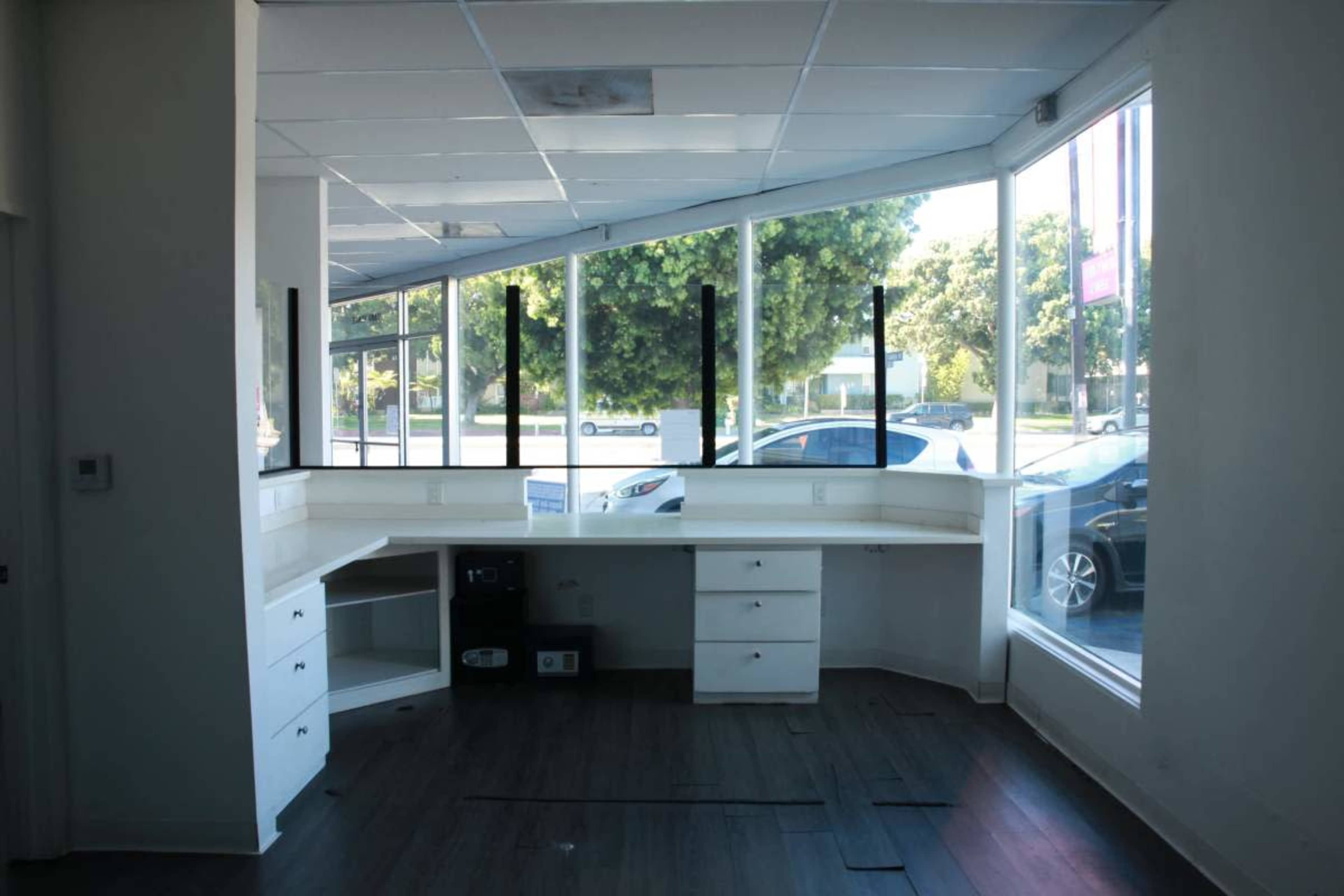 The image shows a vacant office space featuring a white L-shaped desk and large windows that provide a view of the exterior.