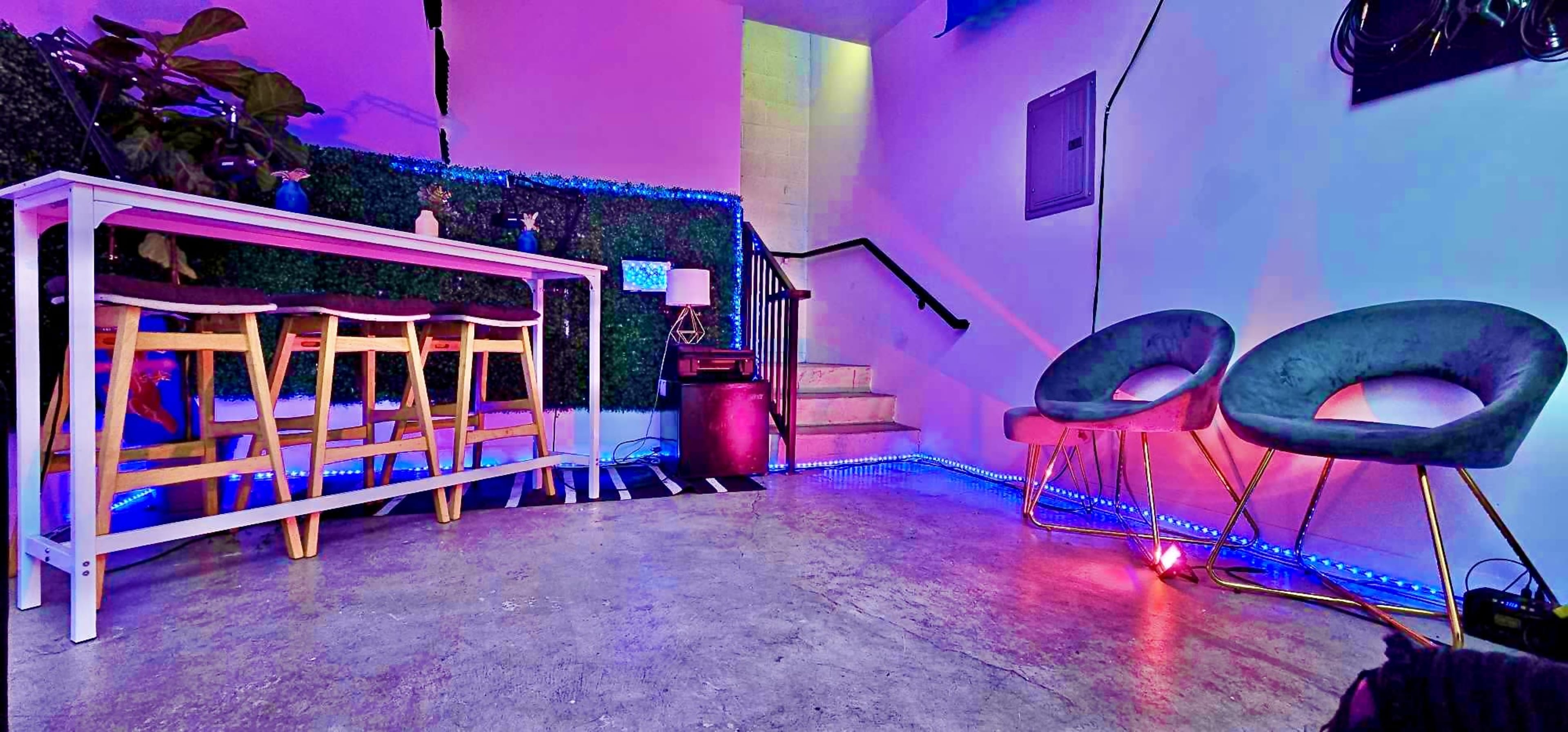 The image shows a modern interior space featuring a white bar table with three wooden stools, two velvet chairs, and a staircase, surrounded by colorful lighting and greenery on the walls.