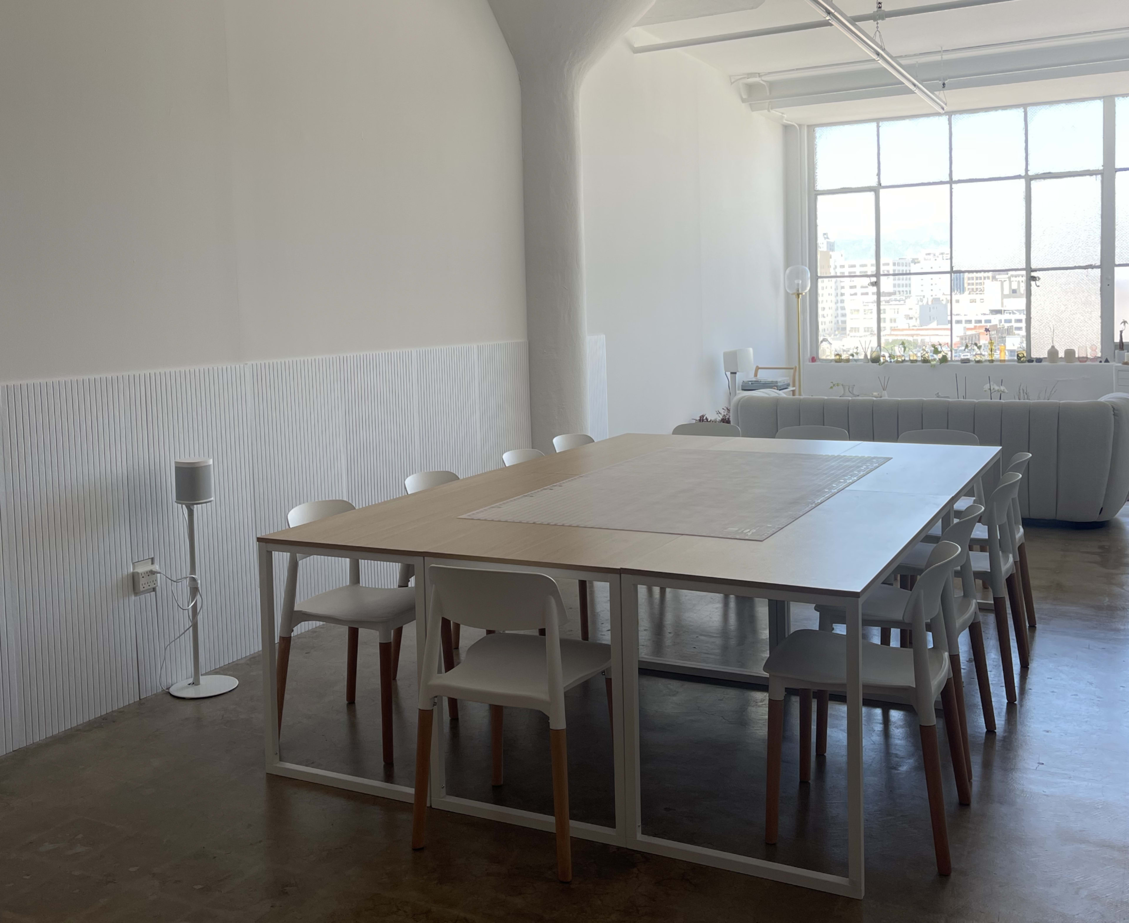 A large, simple dining table is surrounded by white chairs in a bright room with a large window and a couch in the background.
