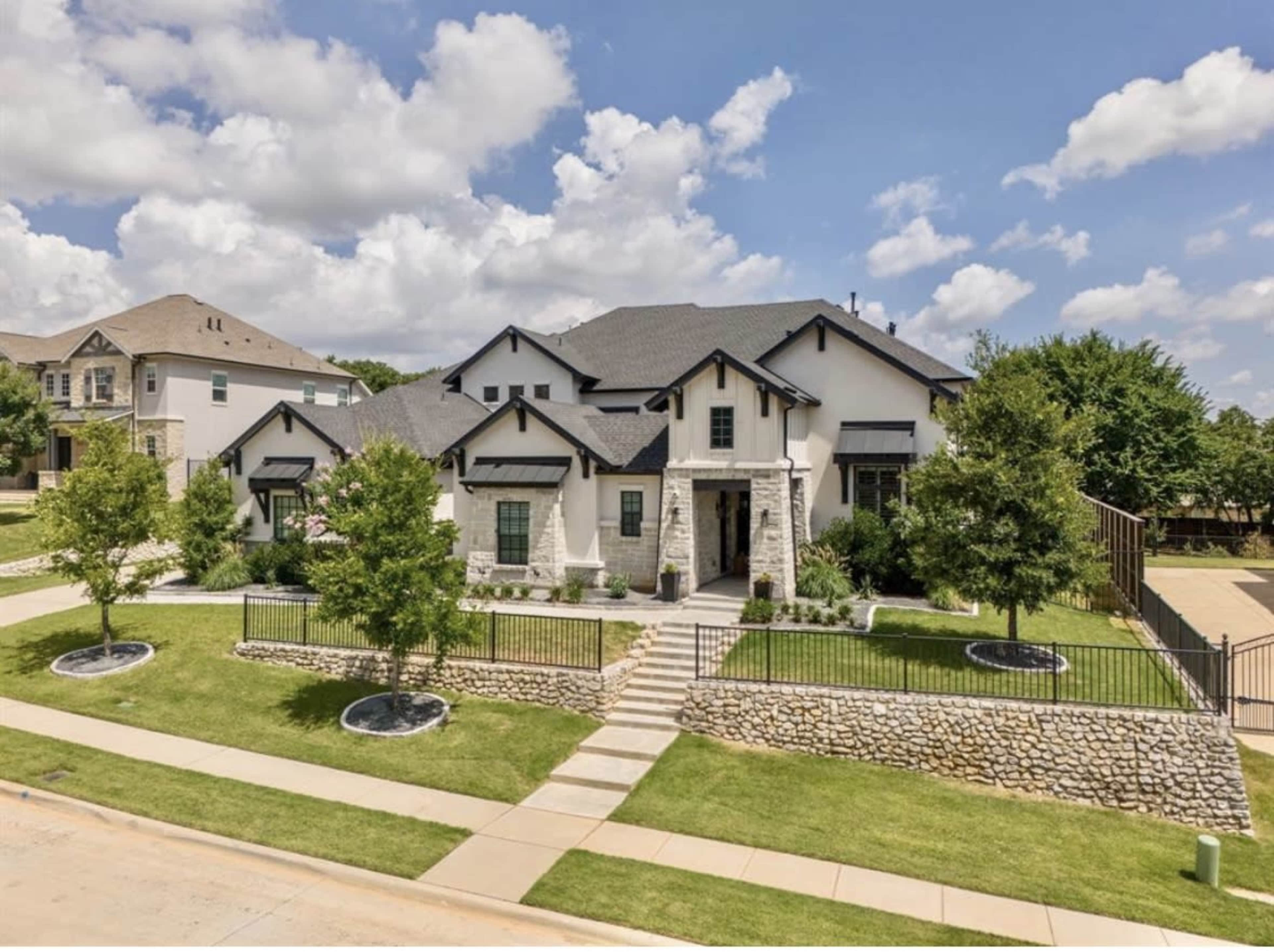 The image shows a large, modern two-story house with a stone facade and a landscaped front yard featuring several trees and a stone retaining wall.