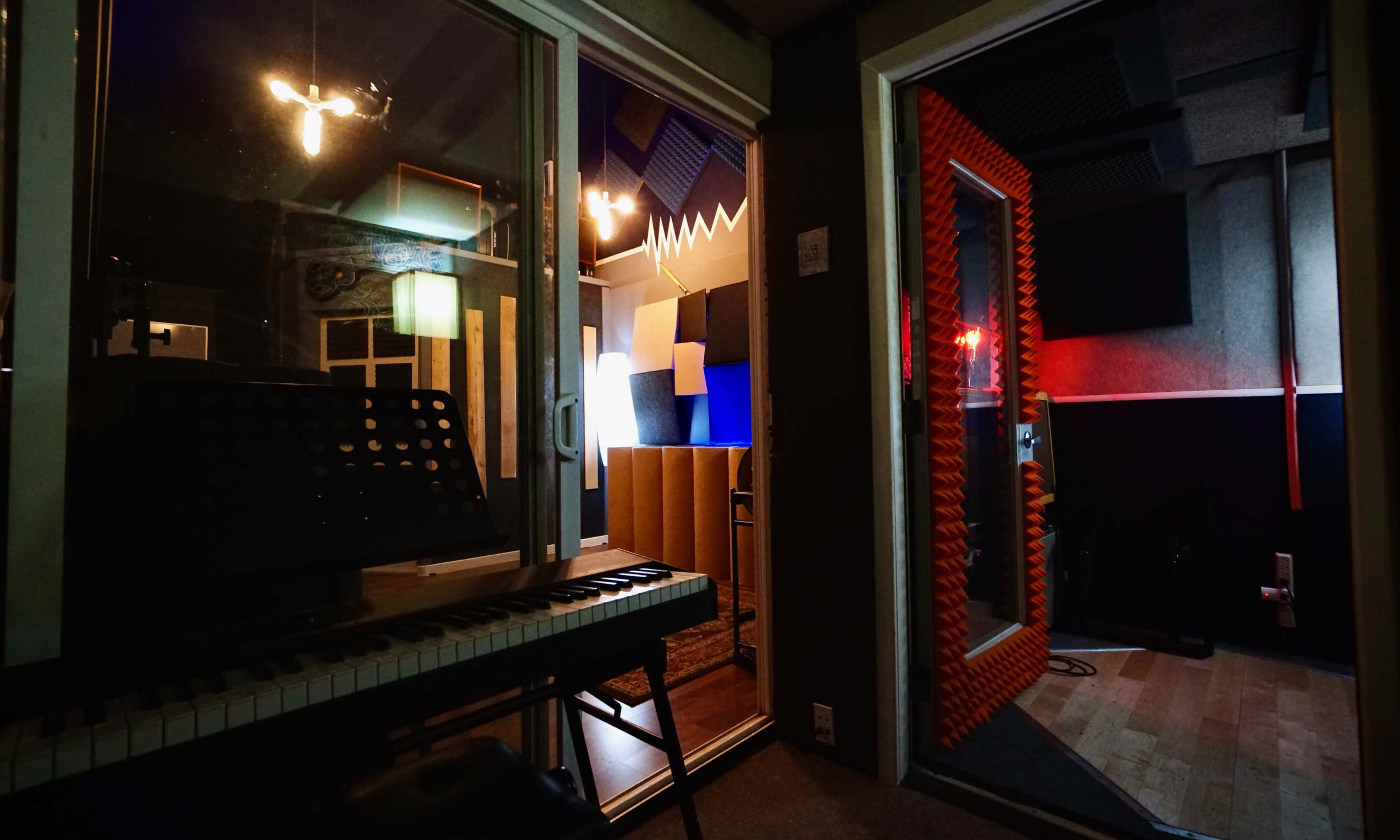 A music recording studio is shown with a keyboard in the foreground and two separate acoustic-treated rooms visible through glass walls.