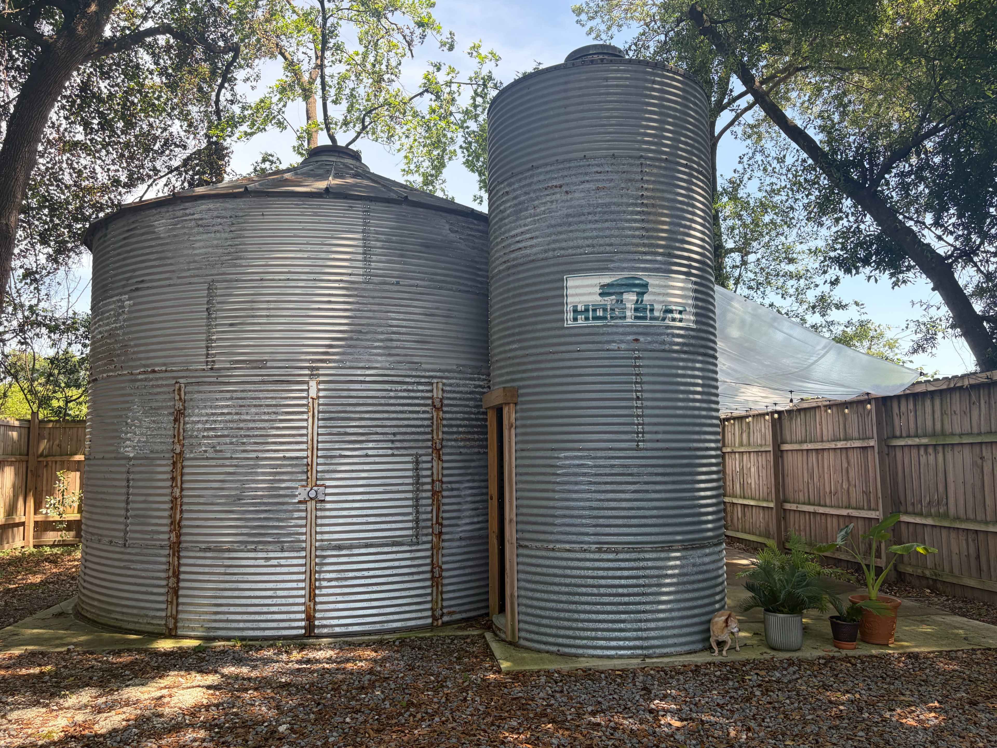 The image shows two weathered metal grain silos beside a wooden fence, surrounded by trees and gravel, with potted plants near the entrance.