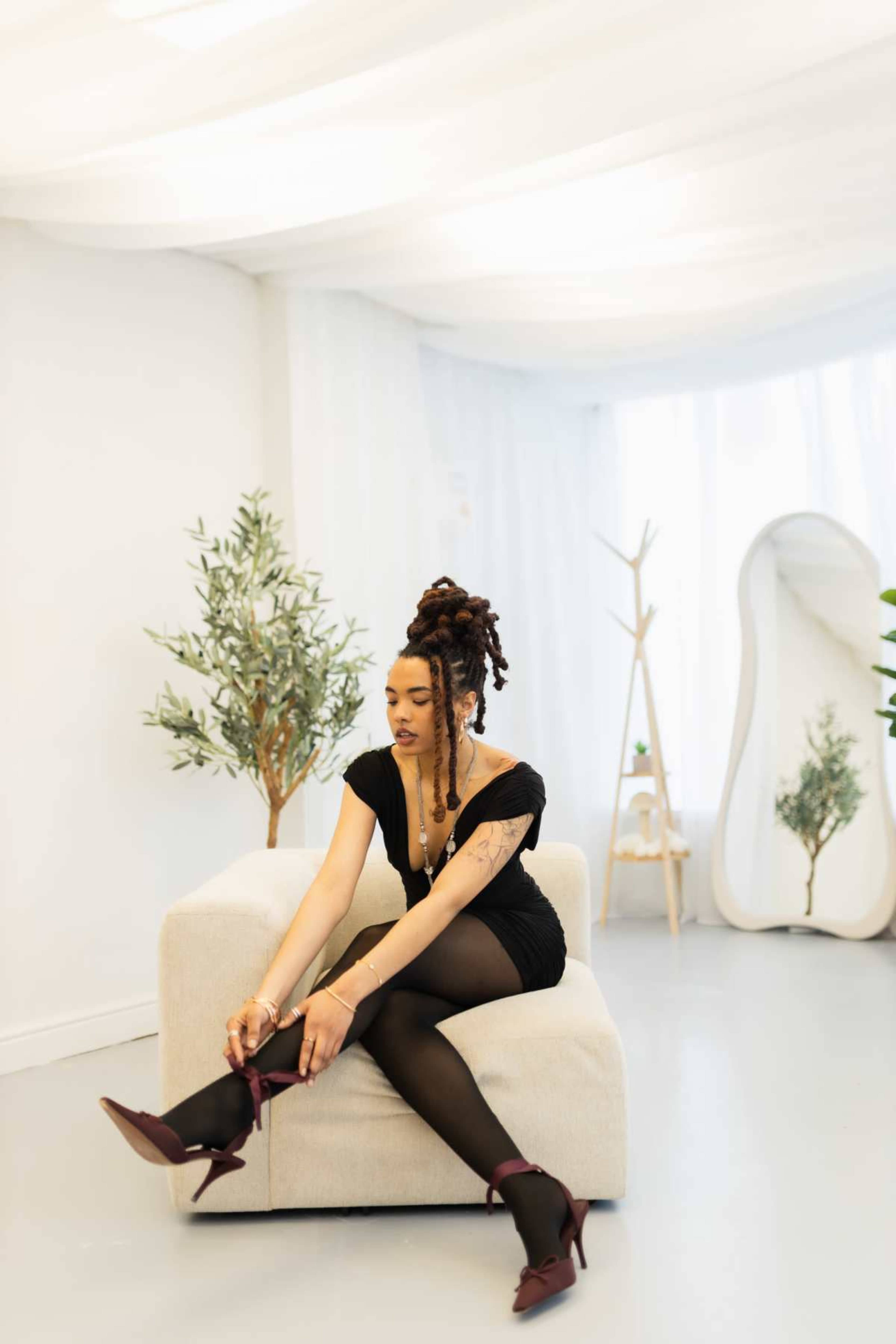 A woman with dreadlocks sits on a light-colored sofa, putting on burgundy high-heeled shoes in a minimalist room with plants and a large mirror.