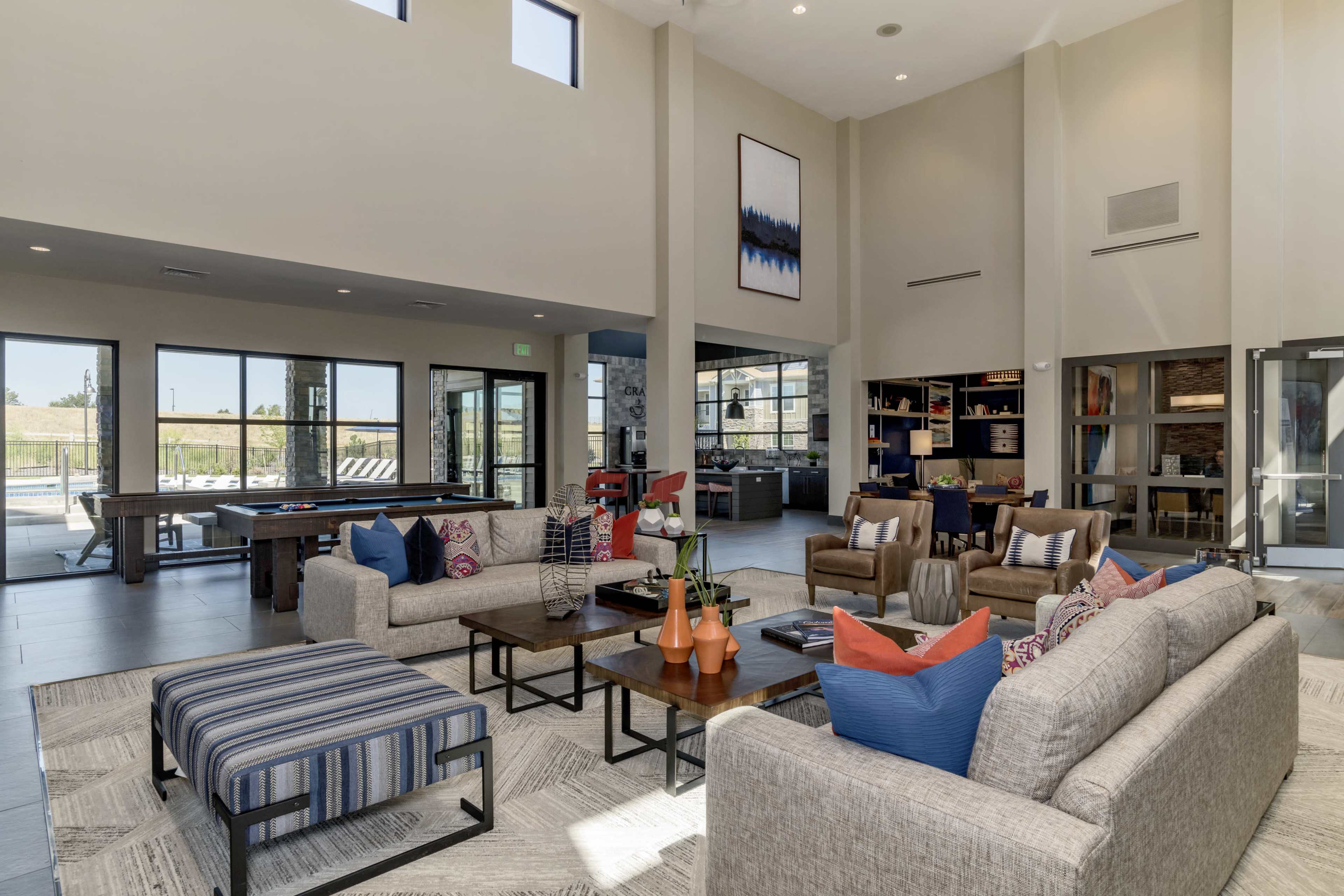 A modern community lounge with a high ceiling, featuring sofas, armchairs, and large windows that provide a view of the outdoors.