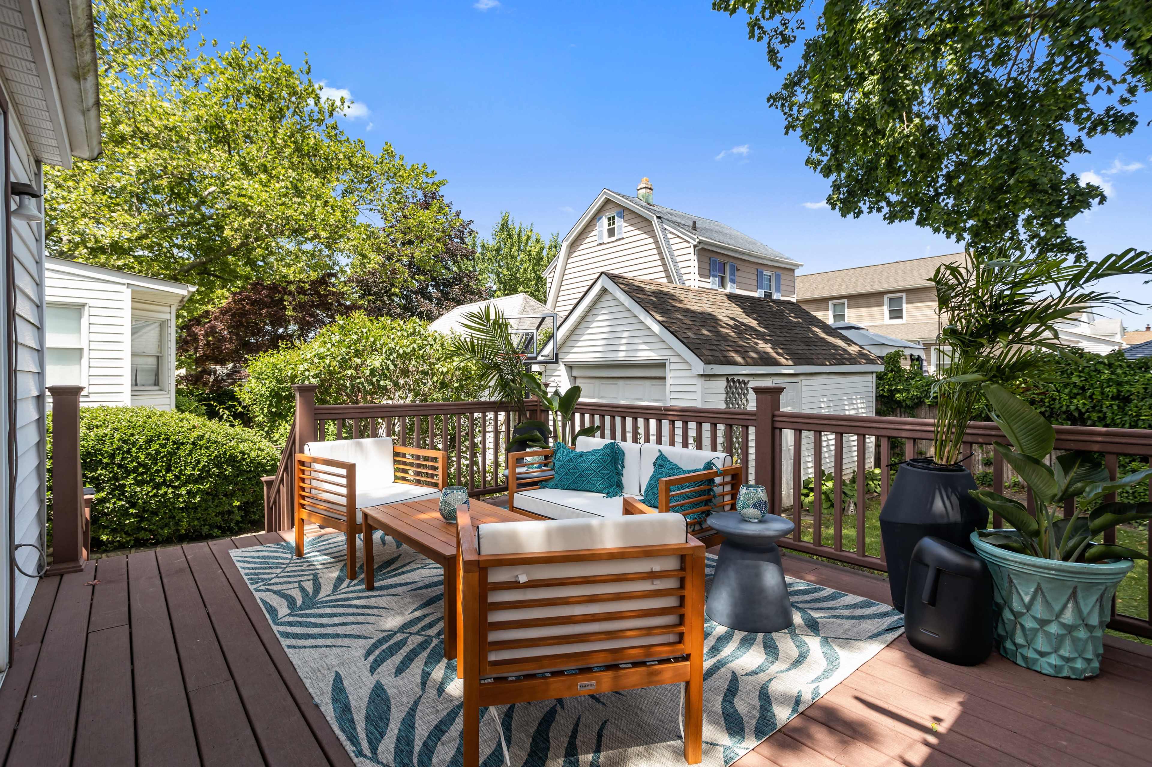 A wooden deck features outdoor seating with cushions, surrounded by greenery and nearby houses.