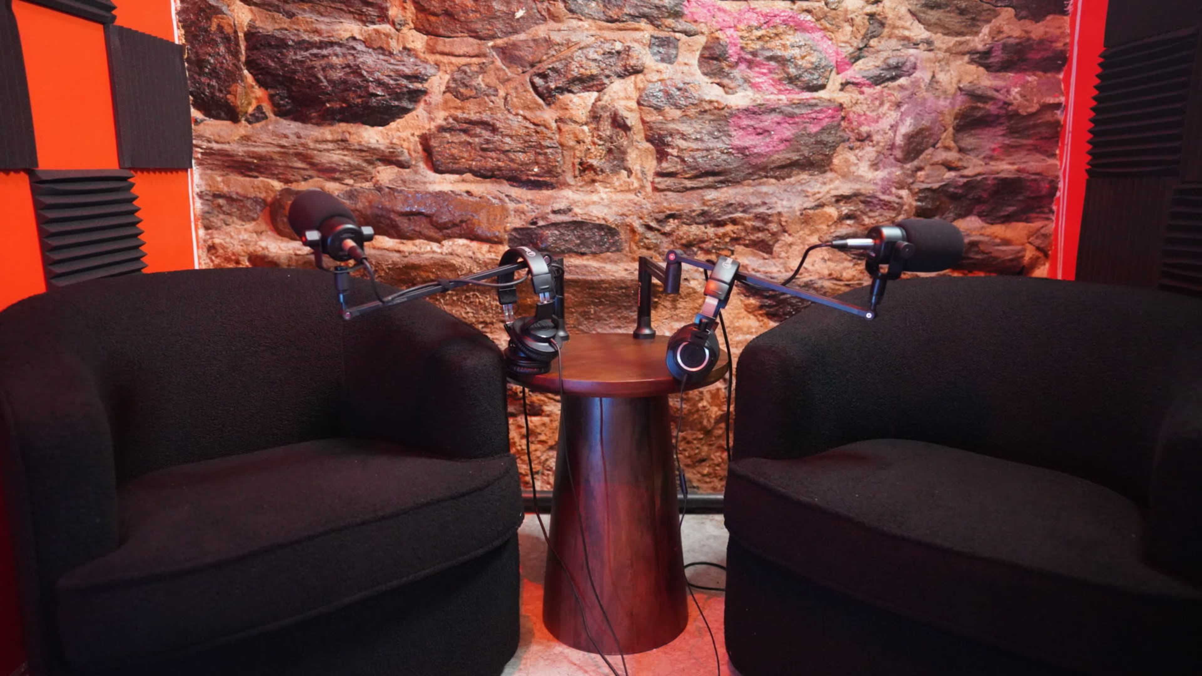 The image shows a recording studio setup with two black chairs, a small wooden table, and microphones mounted on arms against a stone wall.