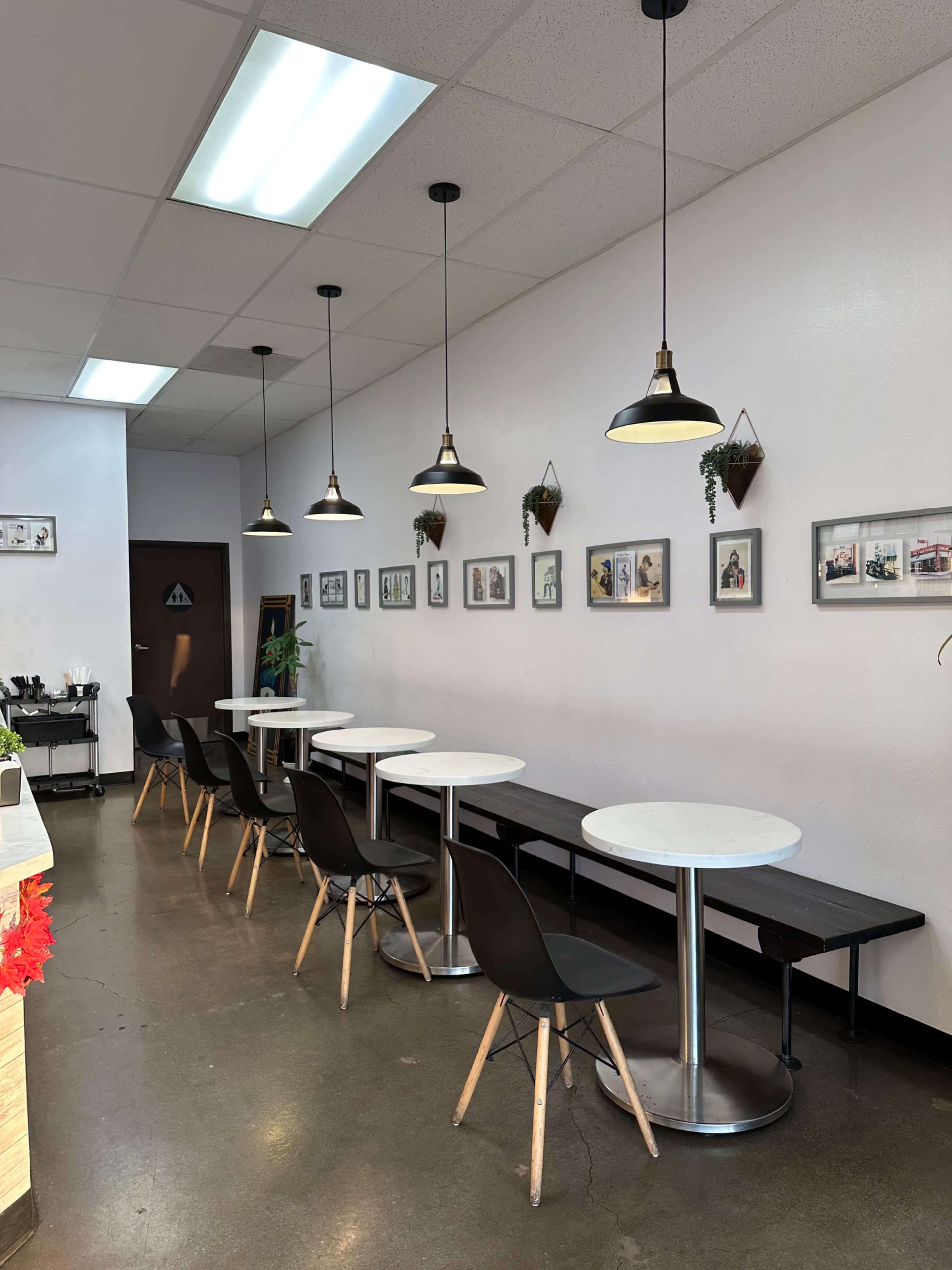 A bright café interior with several white tables, black chairs, and wall-mounted framed photographs.