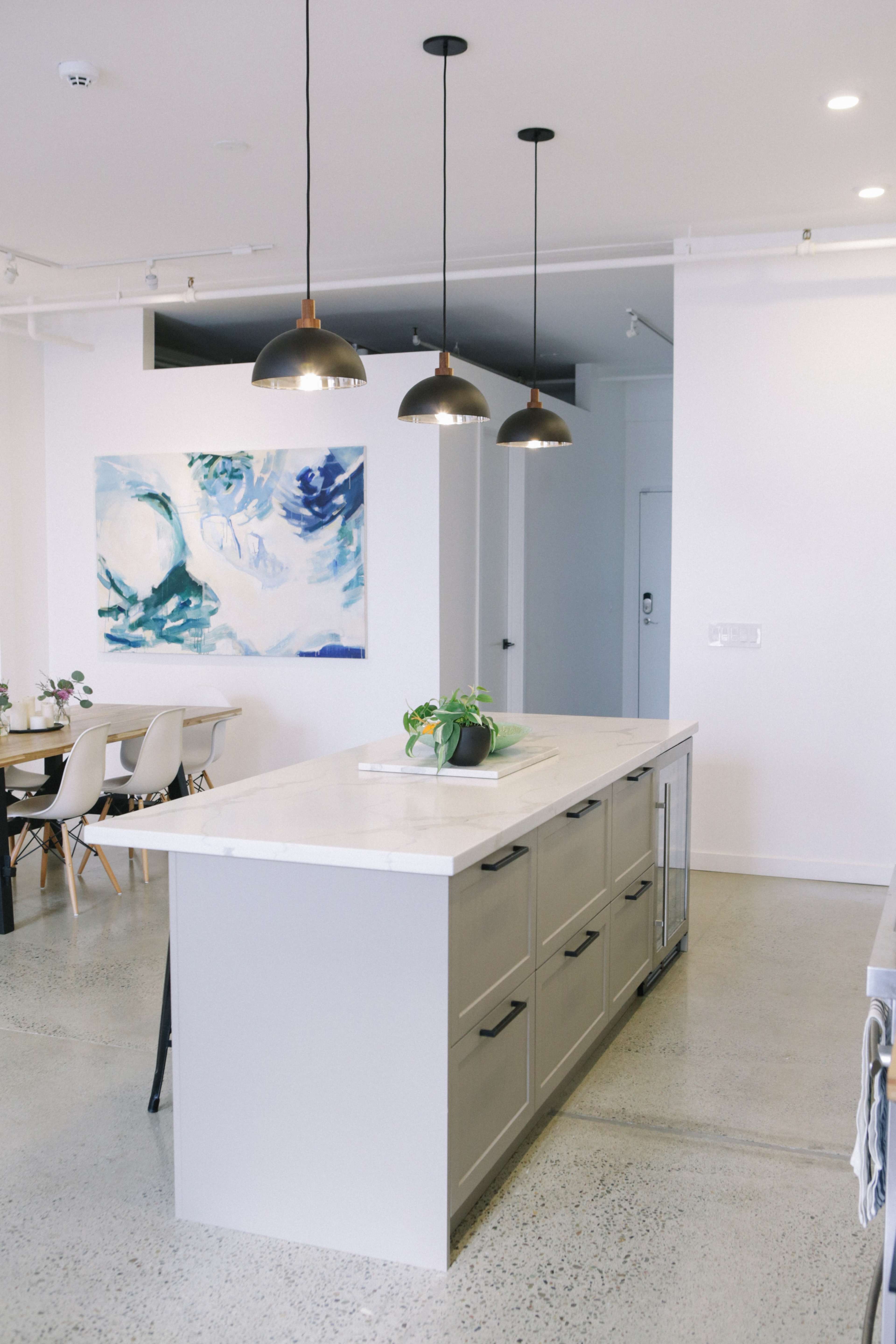 A modern kitchen features a central island with drawers, pendant lights above, and a large abstract painting on the wall.