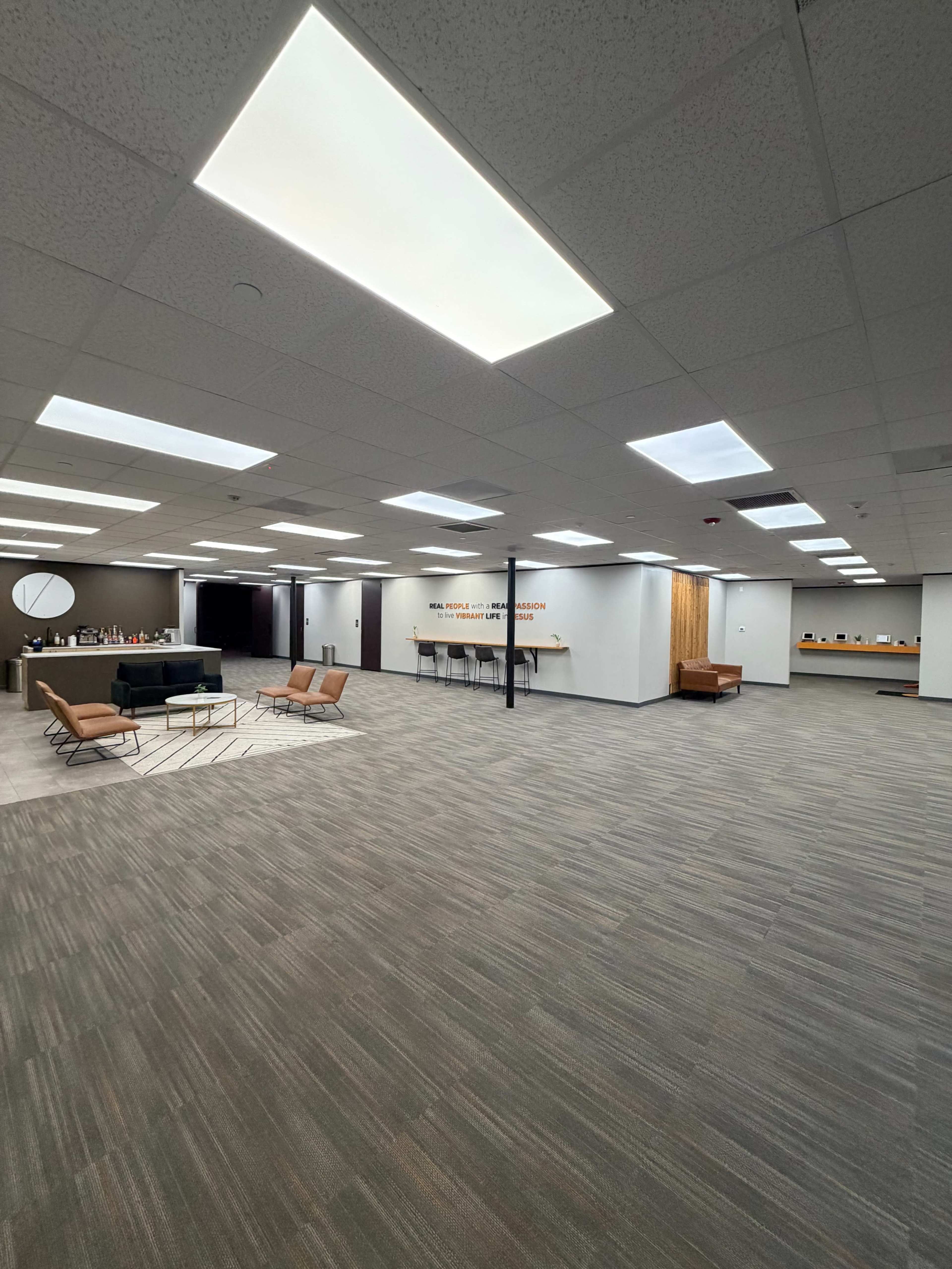 The image shows a spacious, modern office interior with a seating area, a reception desk, and ceiling lights illuminating the gray carpeted floor.