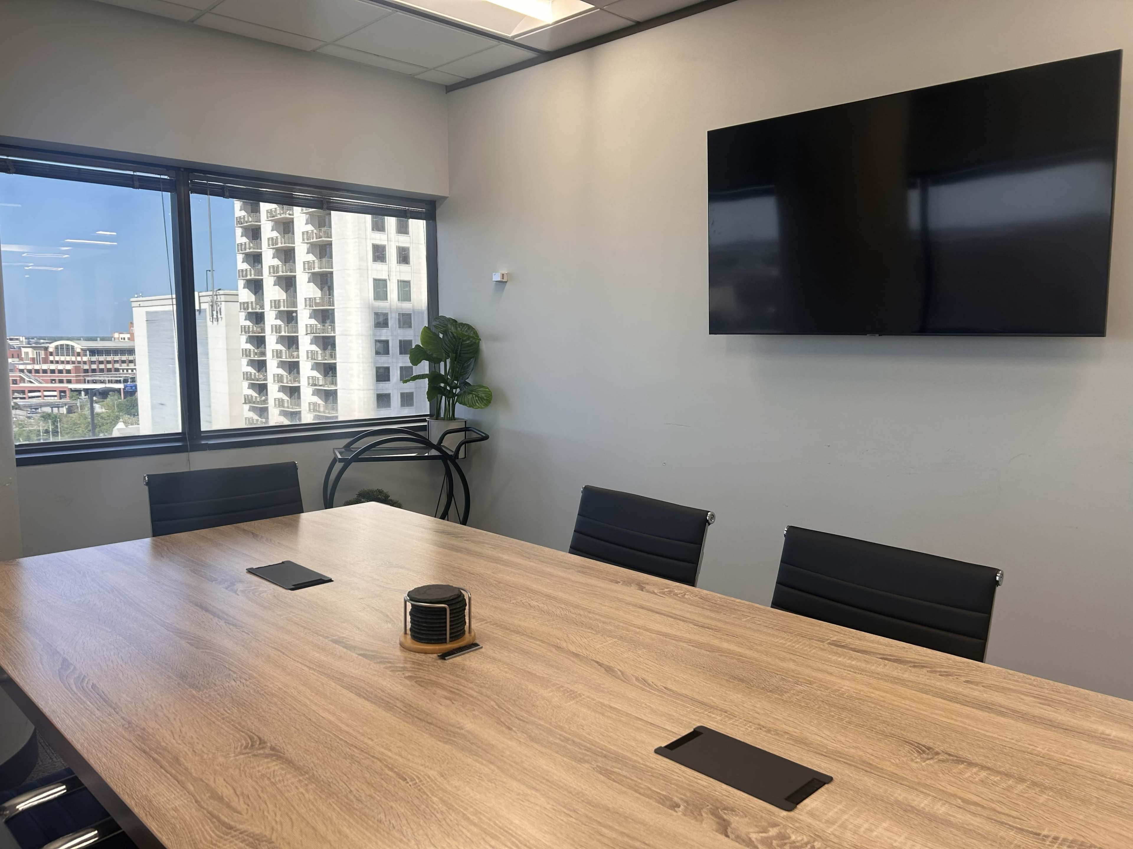 The image shows a modern conference room with a wooden table, black chairs, a large screen on the wall, and a window displaying an urban landscape outside.