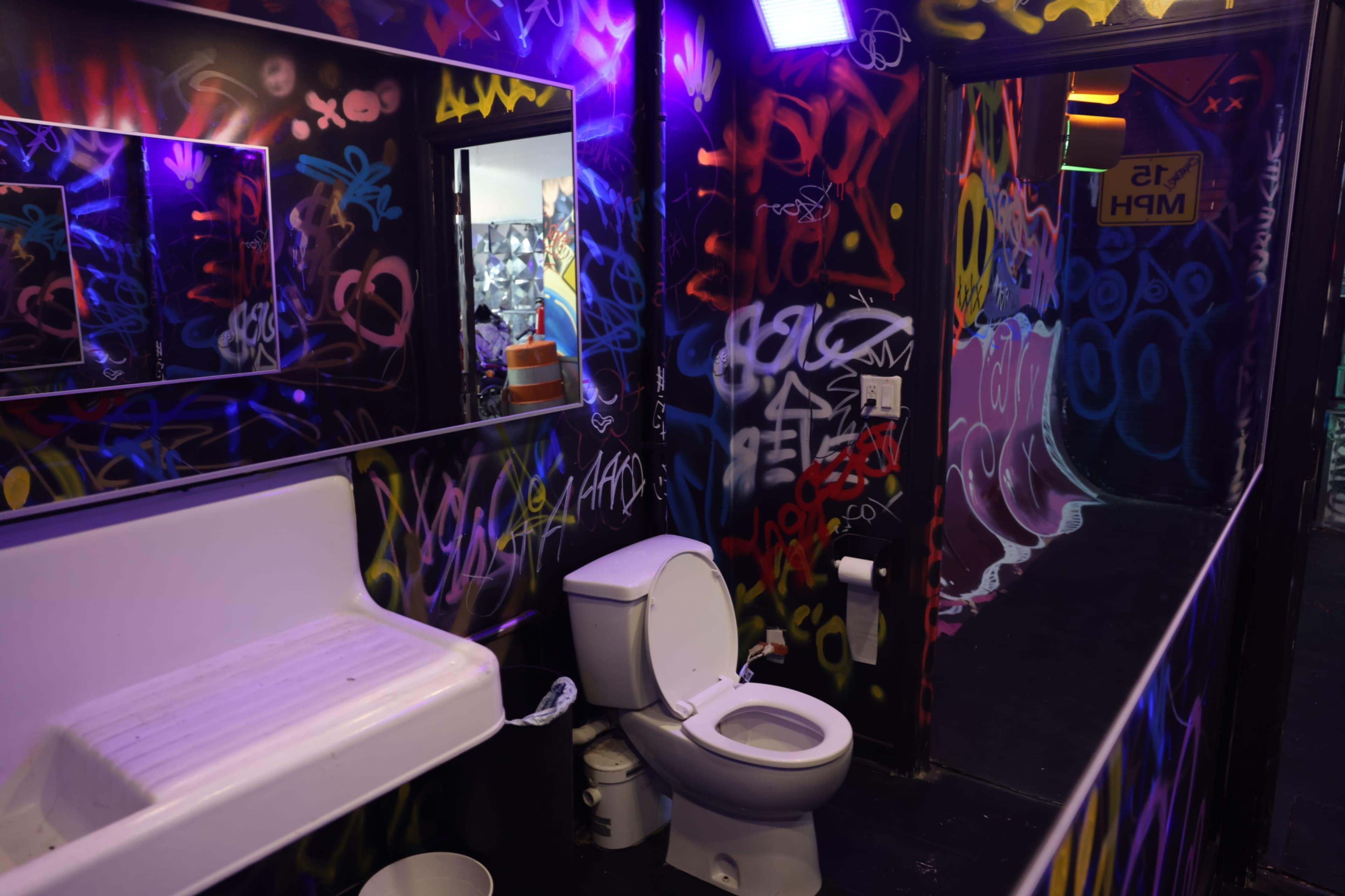 The image shows a graffiti-covered bathroom with a toilet, sink, and mirrors in a dimly lit space.