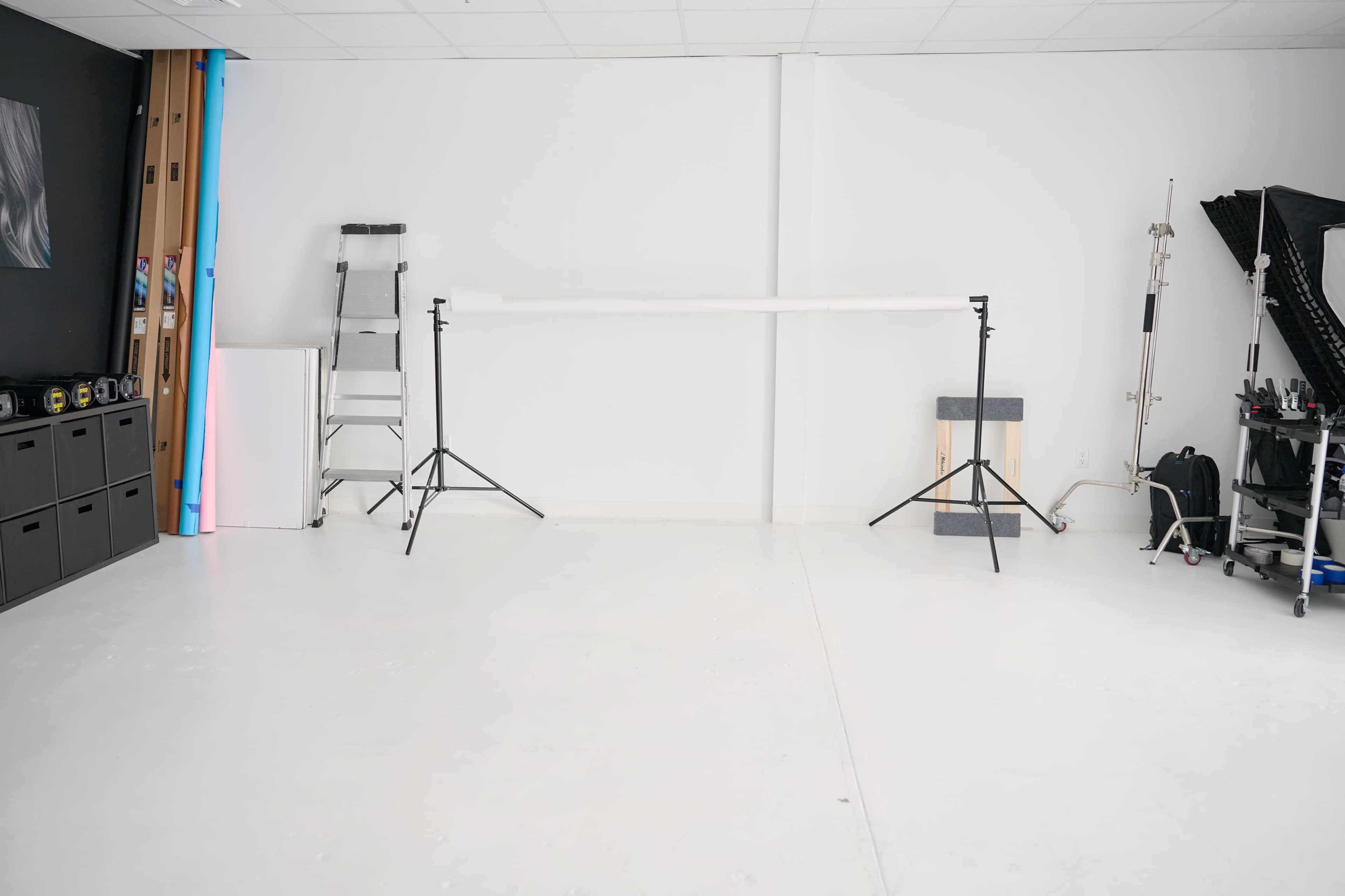 The image shows an empty photography studio with a seamless backdrop setup, a ladder, and various equipment organized along the walls.