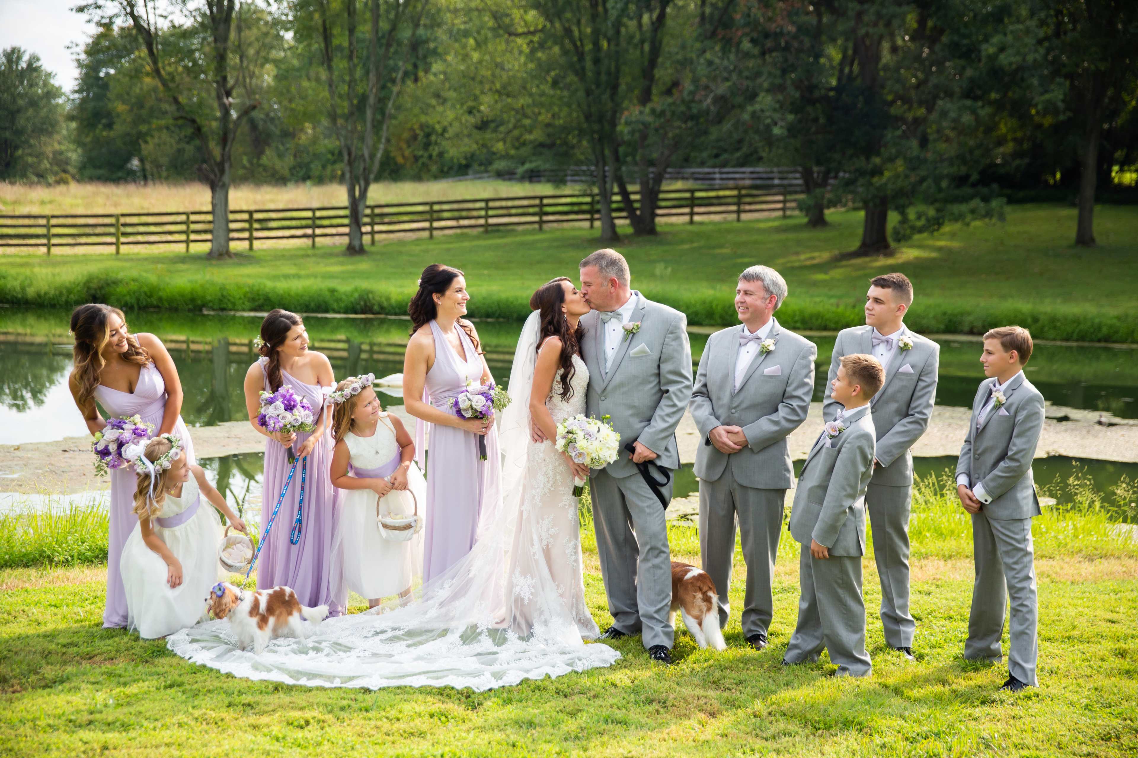 A bridal party stands by a pond, with the bride and groom sharing a kiss at the center, surrounded by bridesmaids, groomsmen, and a few children.